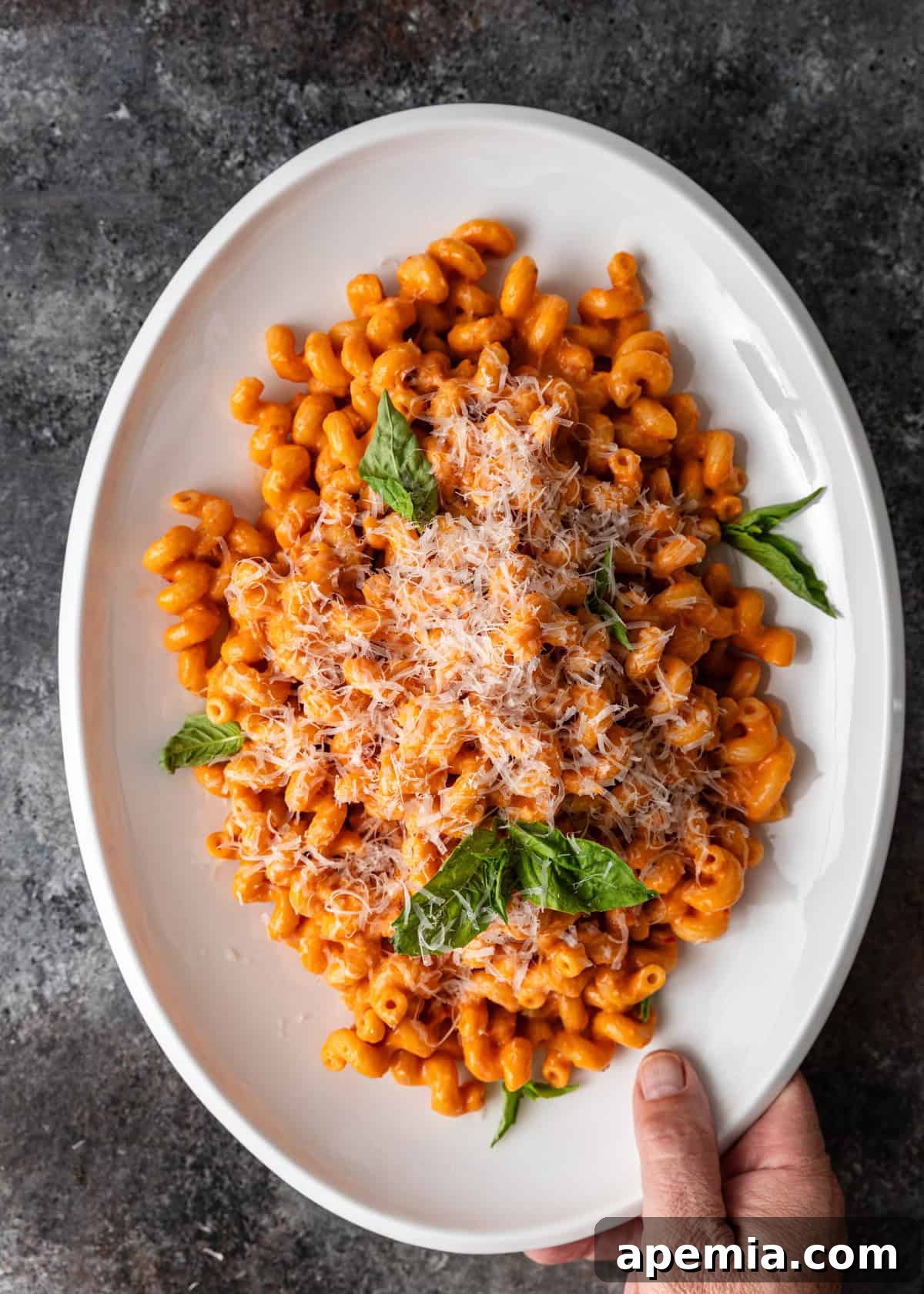 Overhead view of a hand holding a platter of tomato confit pasta, generously topped with grated parmesan cheese