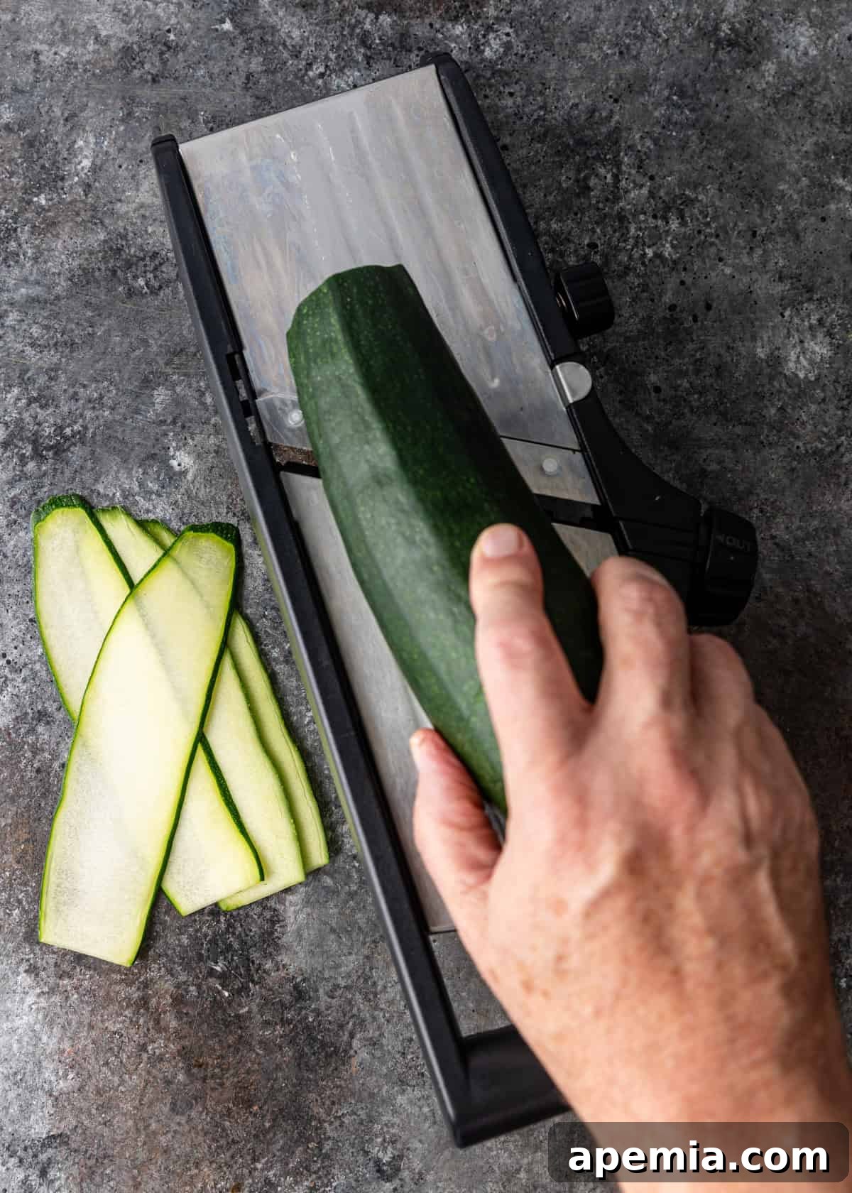 Overhead process shot: Thinly peeling zucchini into long ribbons using a vegetable mandoline for consistent slices.