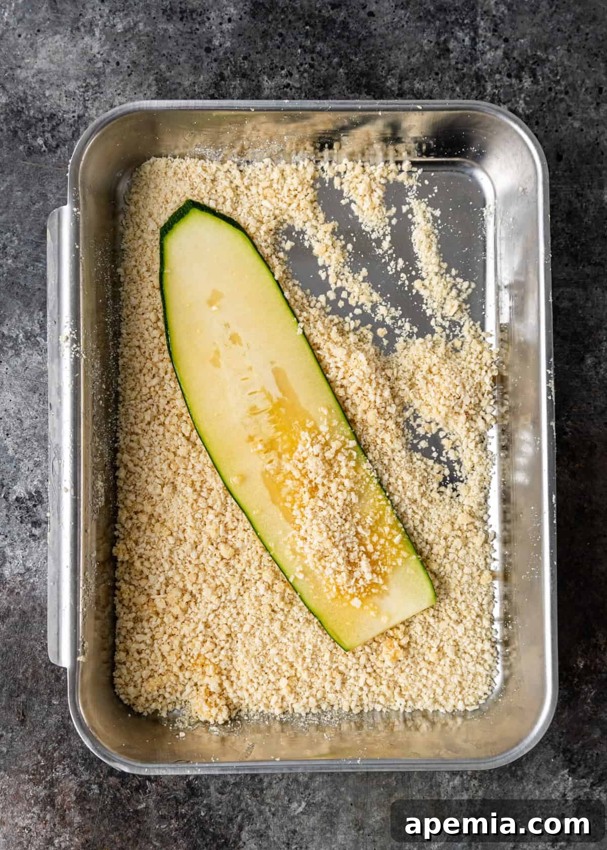 Overhead process shot: A zucchini ribbon being dipped and coated in a mixture of breadcrumbs and grated Parmesan cheese.