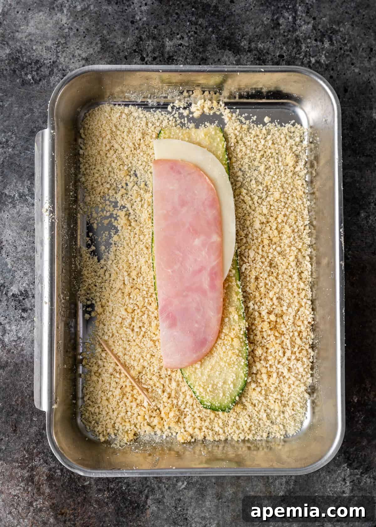 Overhead process shot: A breaded zucchini ribbon topped with a slice of provolone cheese and a slice of ham or prosciutto, ready for rolling.