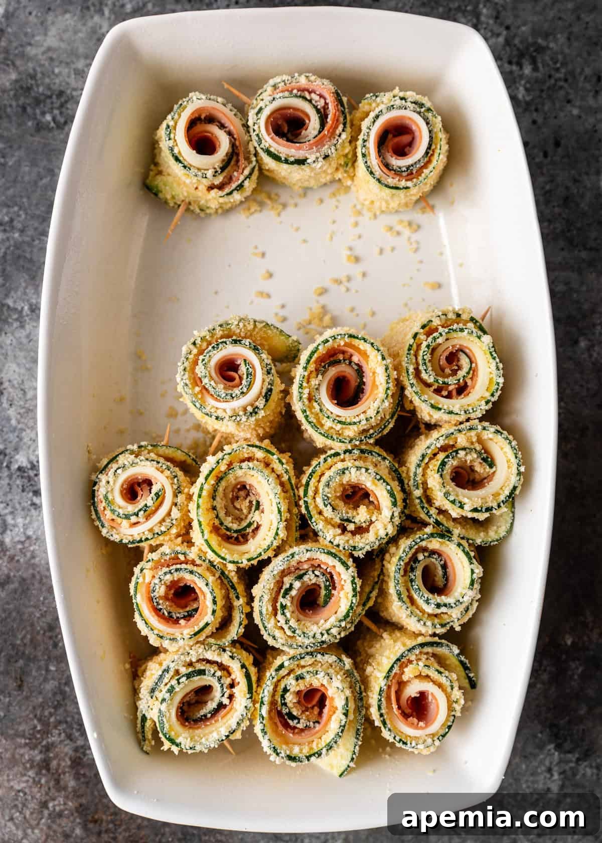 Overhead process shot: Several zucchini involtini neatly rolled up and secured with toothpicks, placed in a baking dish, ready for the oven.