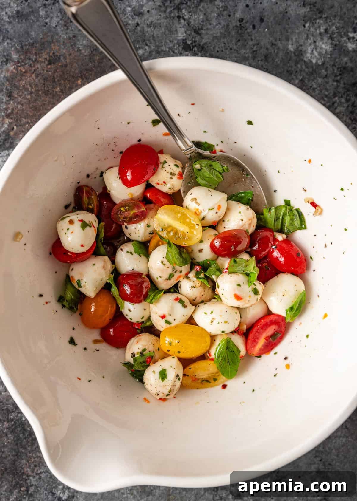 A white mixing bowl filled with fresh mozzarella balls, vibrant cherry tomatoes, and aromatic basil leaves, ready for preparing a Caprese salad.