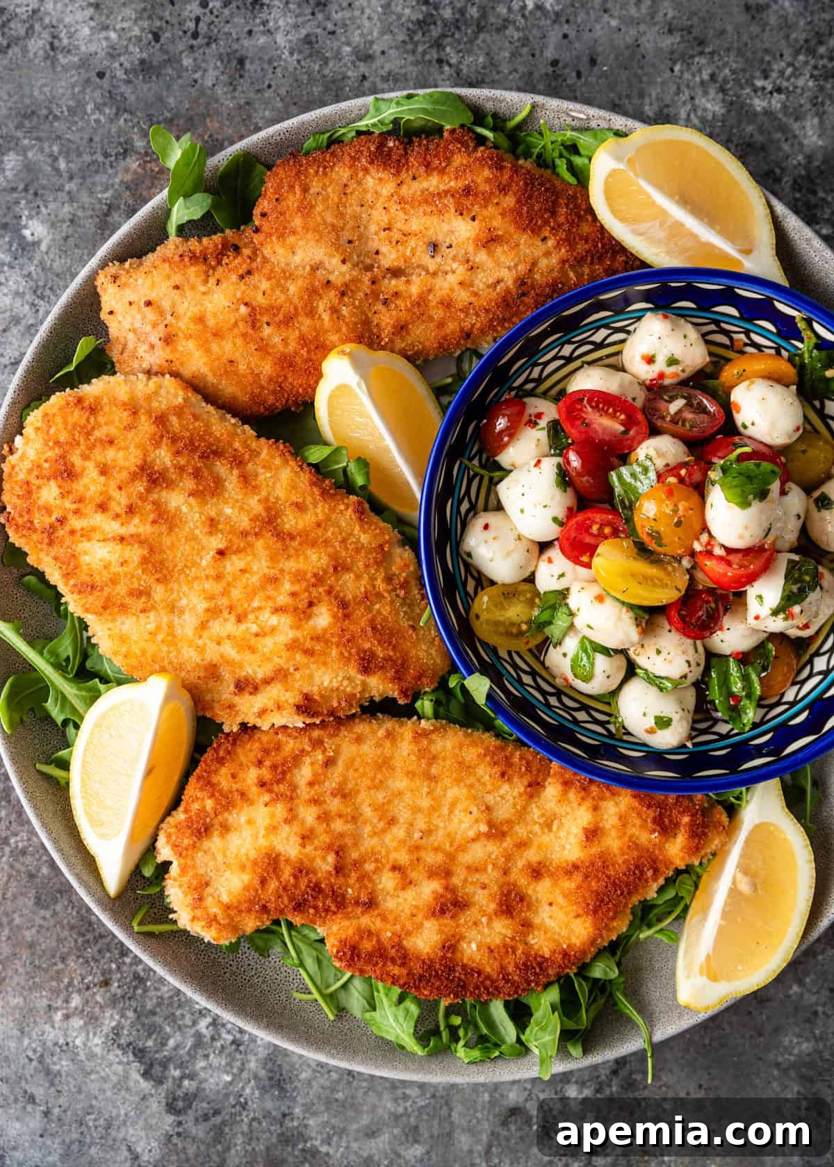 A beautifully plated crispy chicken Milanese recipe over fresh greens, garnished with lemon wedges, and accompanied by a colorful Caprese salad, ready to be served.