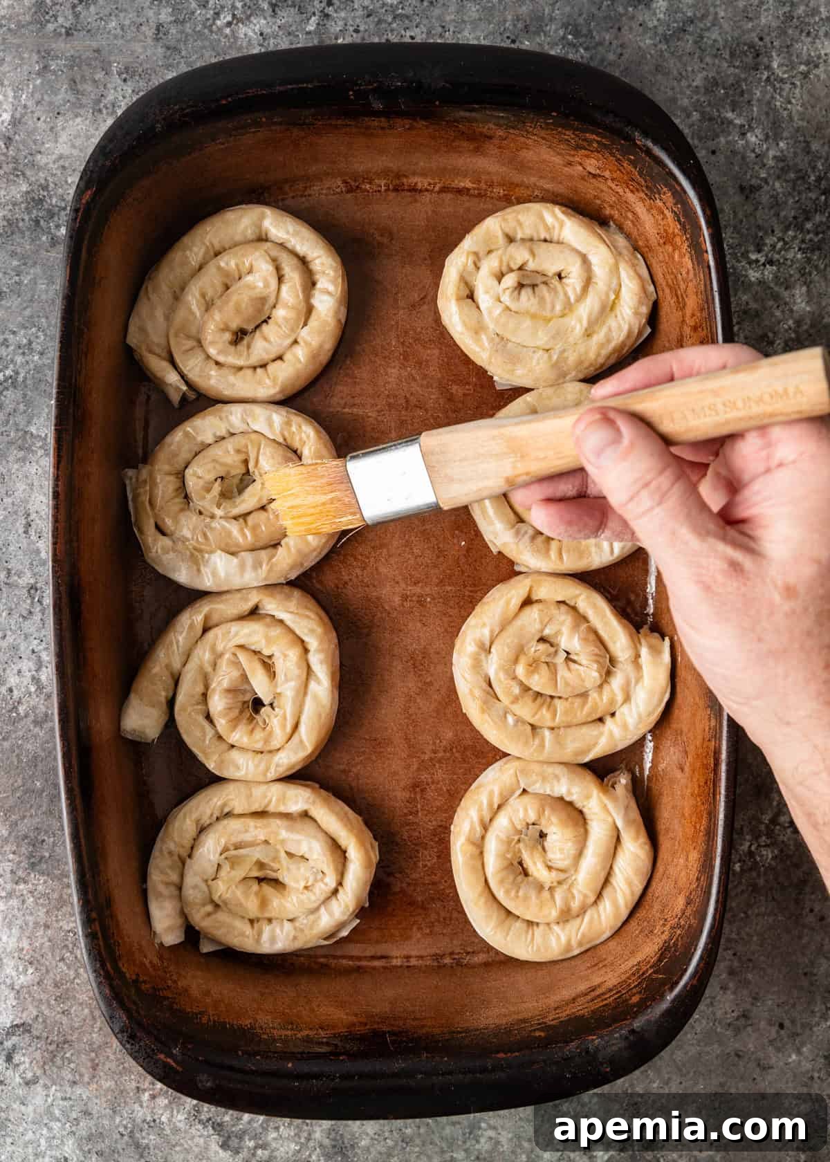 Mhencha: Moroccan Coiled Almond Delight 11 Overhead process shot: brushing extra melted butter over uncooked coiled Mhencha pastries in a baking dish.