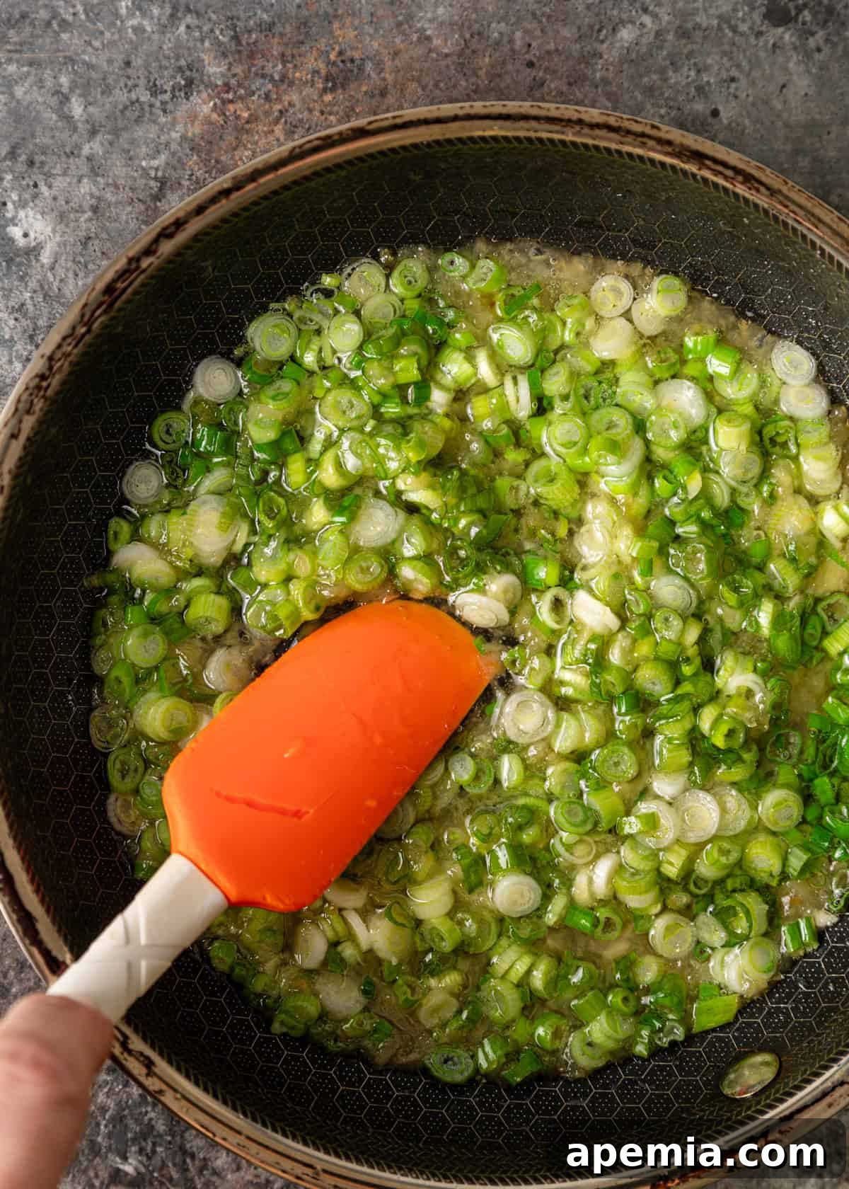 Overhead process shot: an orange spatula stirring ginger scallion sauce as it cooks gently in a small skillet
