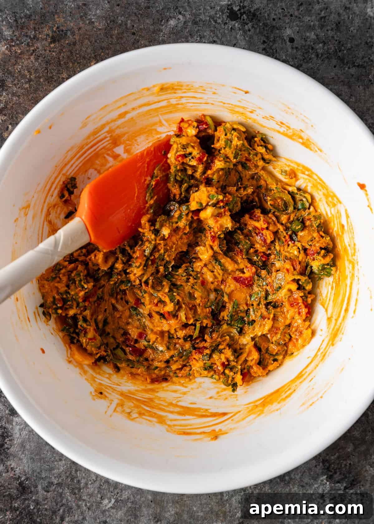 Overhead process shot showing the herb chili butter being combined with a red spatula, creating a vibrant, uniform mixture.