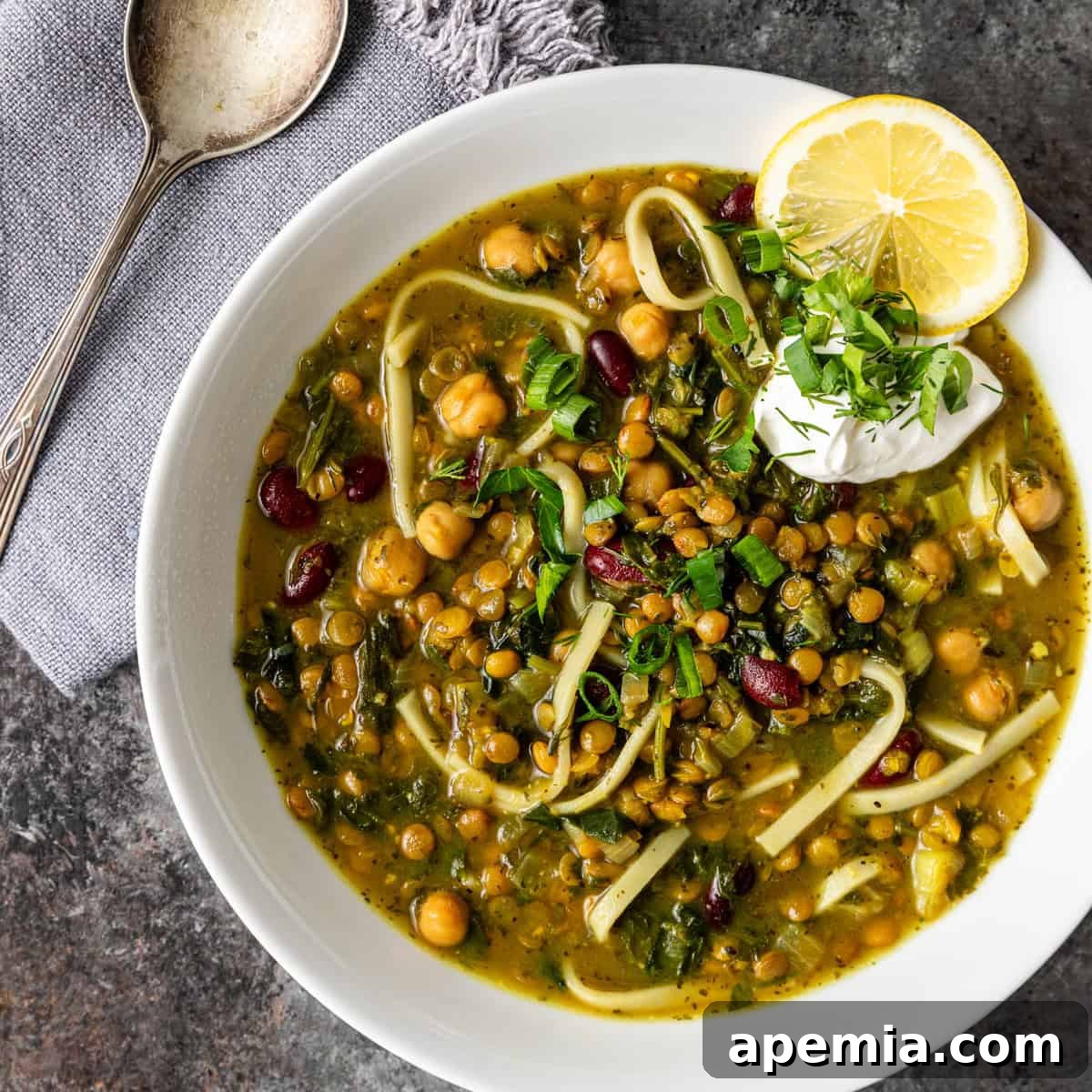 Overhead view of a large bowl of Ash Reshteh noodle soup, garnished with chickpeas, a lemon wedge, and fresh cheese.