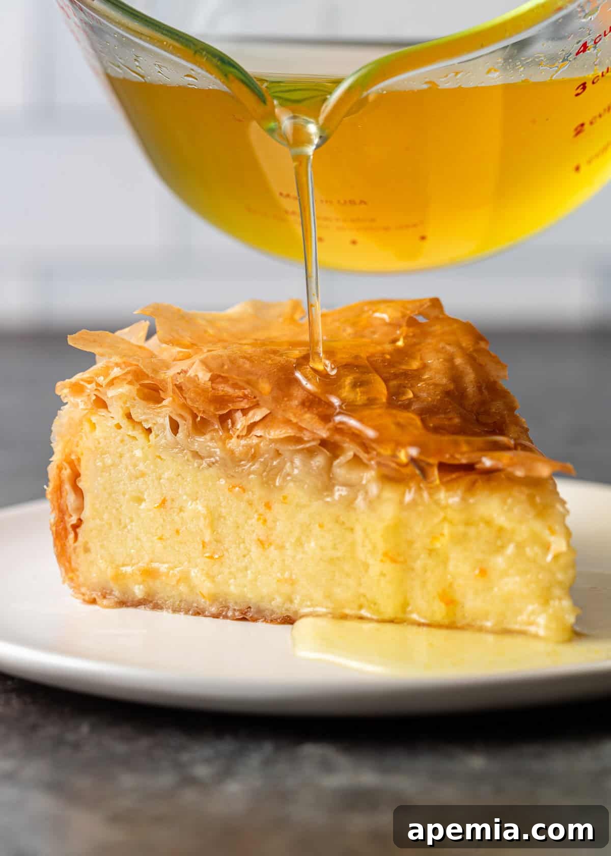 Side view of a hand pouring a golden stream of honey over a delectable slice of Greek filo custard pie, highlighting the rich syrup.