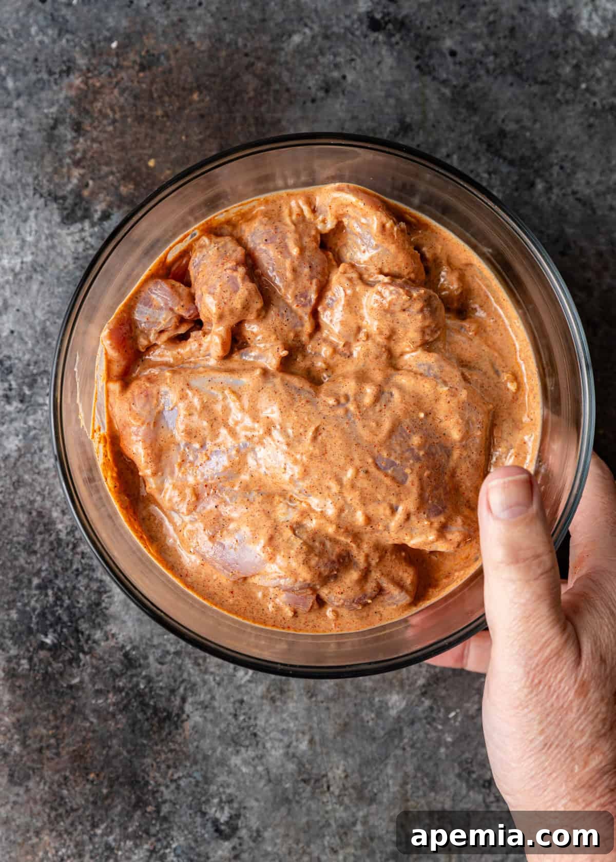 Overhead shot of a hand holding a glass bowl filled with chicken pieces marinating in a vibrant red mixture, ready for cooking.