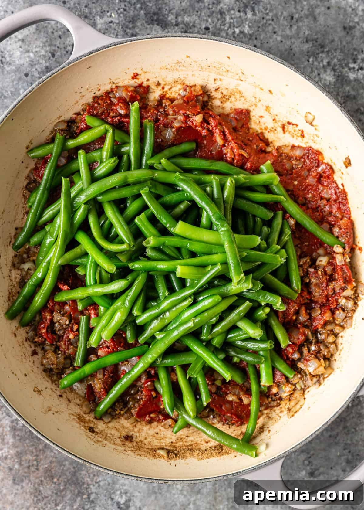 An overhead process shot of fresh green beans being added to a simmering skillet, joining the fragrant sautéed onions, spices, and tomato paste, ready to absorb all the delicious flavors.