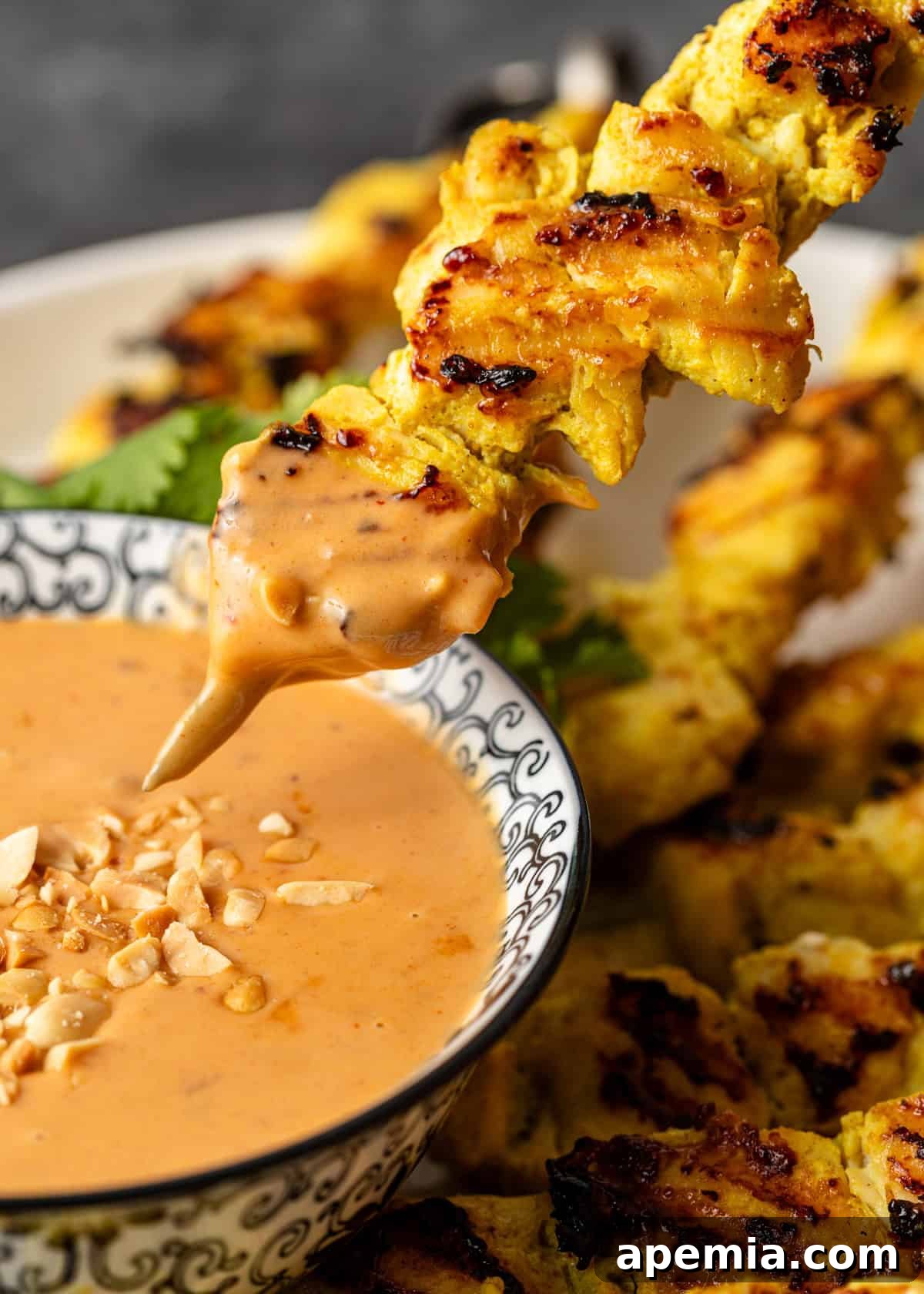 A close-up, side view captures a delicious chicken skewer being dipped into a luscious bowl of homemade Thai peanut sauce, highlighting its creamy texture.