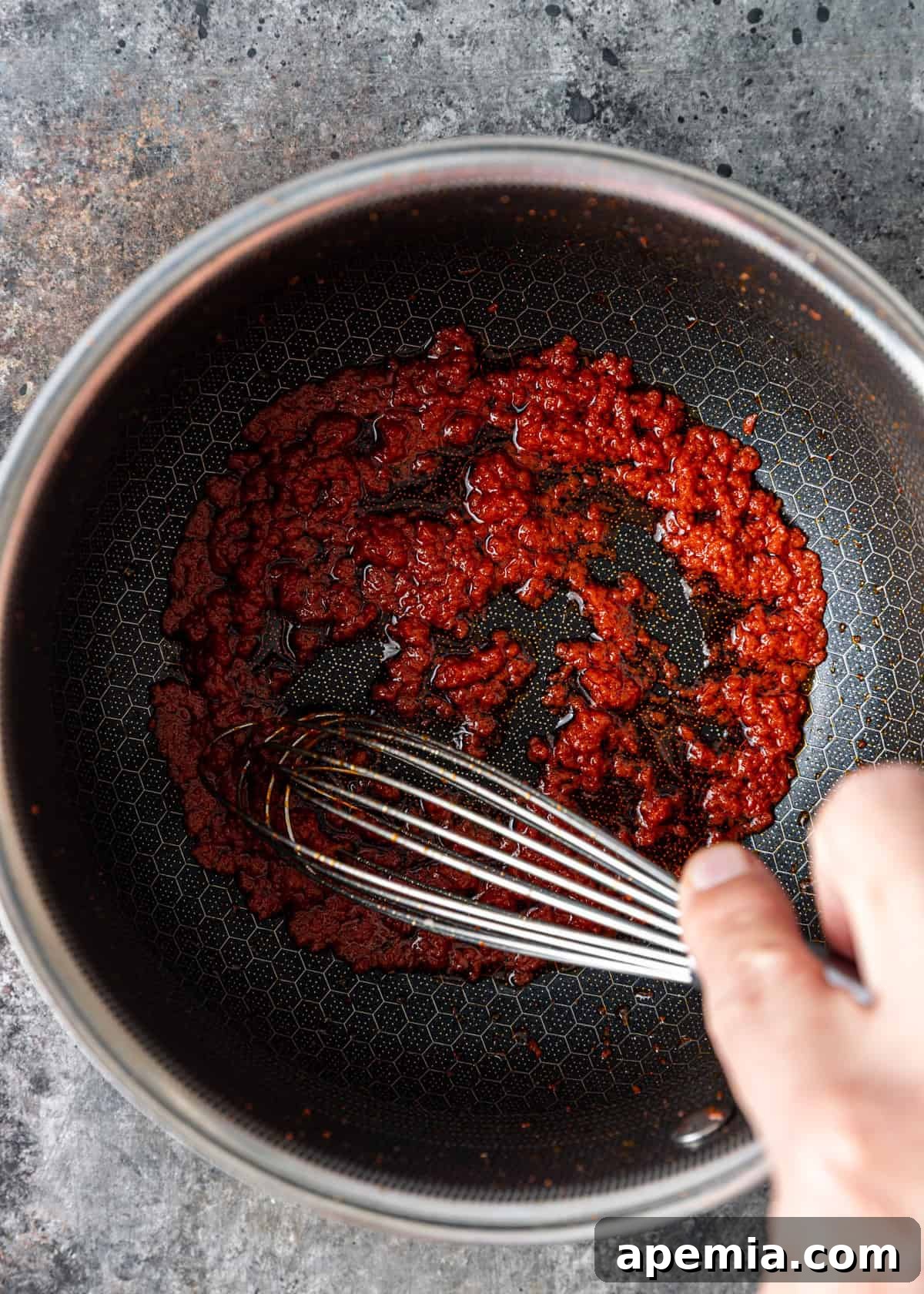 An overhead shot showing the process of heating and stirring vibrant Thai red curry paste in a skillet with a whisk, ensuring all the aromatic spices are bloomed perfectly.