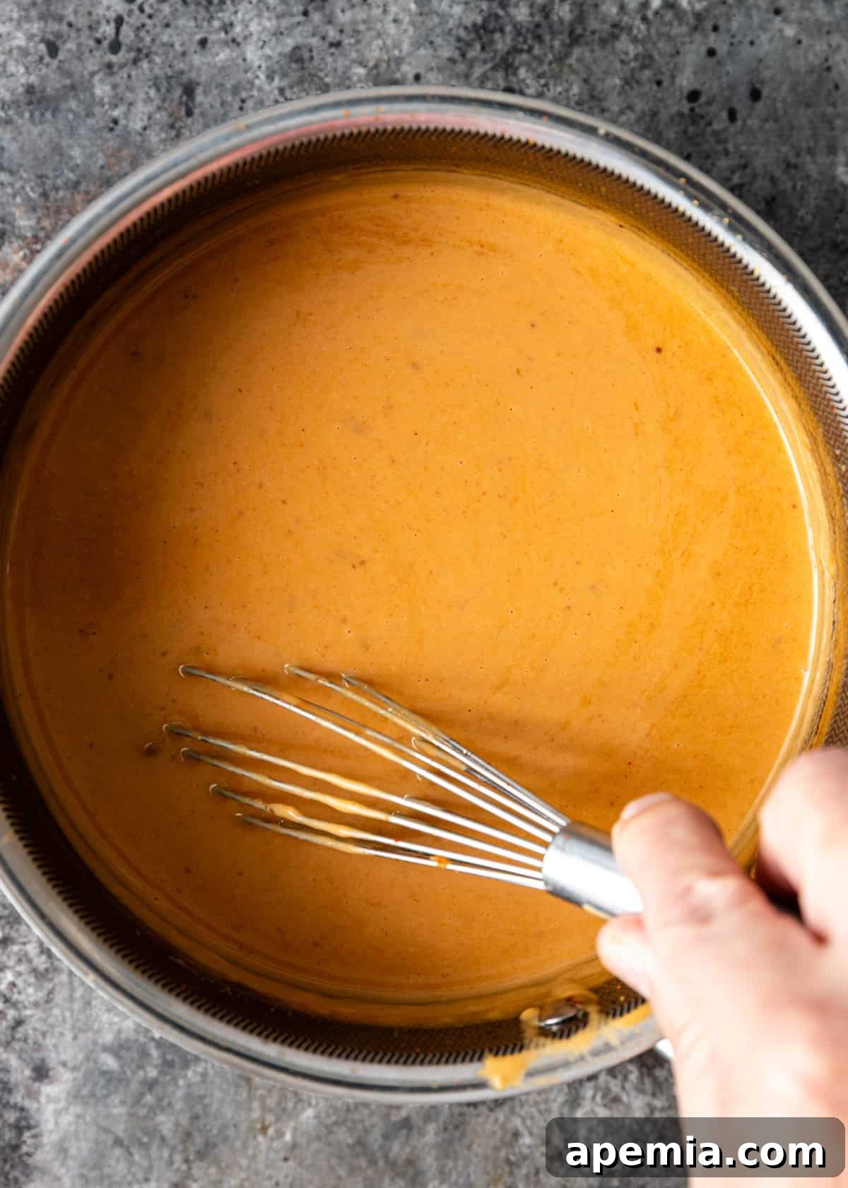 An overhead view captures a whisk diligently stirring the creamy, rich Thai peanut sauce in a bowl, showcasing its smooth and inviting texture.