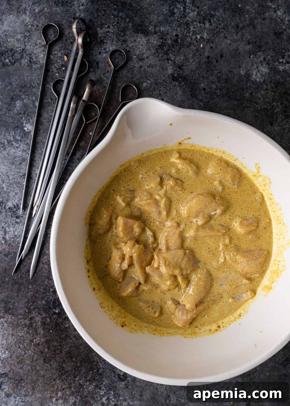 Overhead shot of raw chicken pieces marinating in a rich, creamy sauce inside a bowl, ready to be threaded onto skewers for satay.