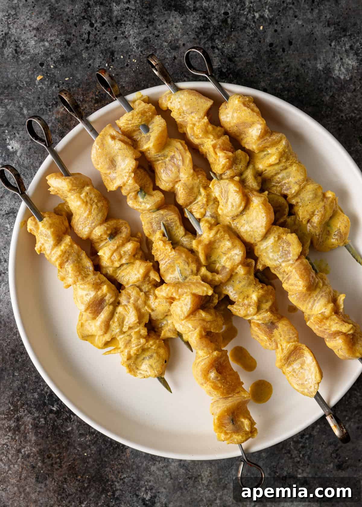 Overhead view of perfectly marinated chicken satay skewers arranged on a pristine white plate, gleaming and ready for the grill.