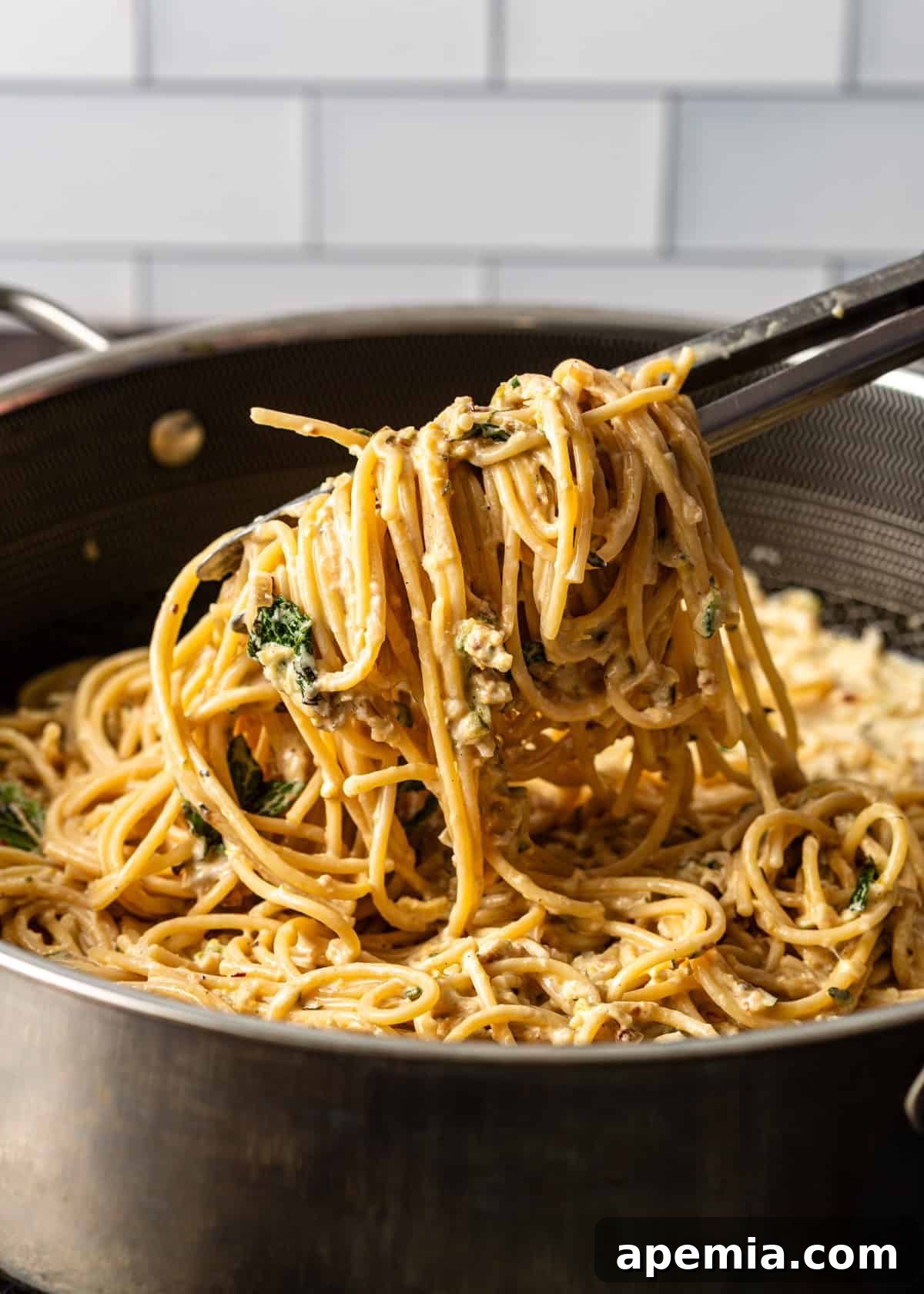 Side view closeup: kitchen tongs lifting a portion of creamy lemon zucchini pasta out of a large skillet, showing the sauce coating the spaghetti