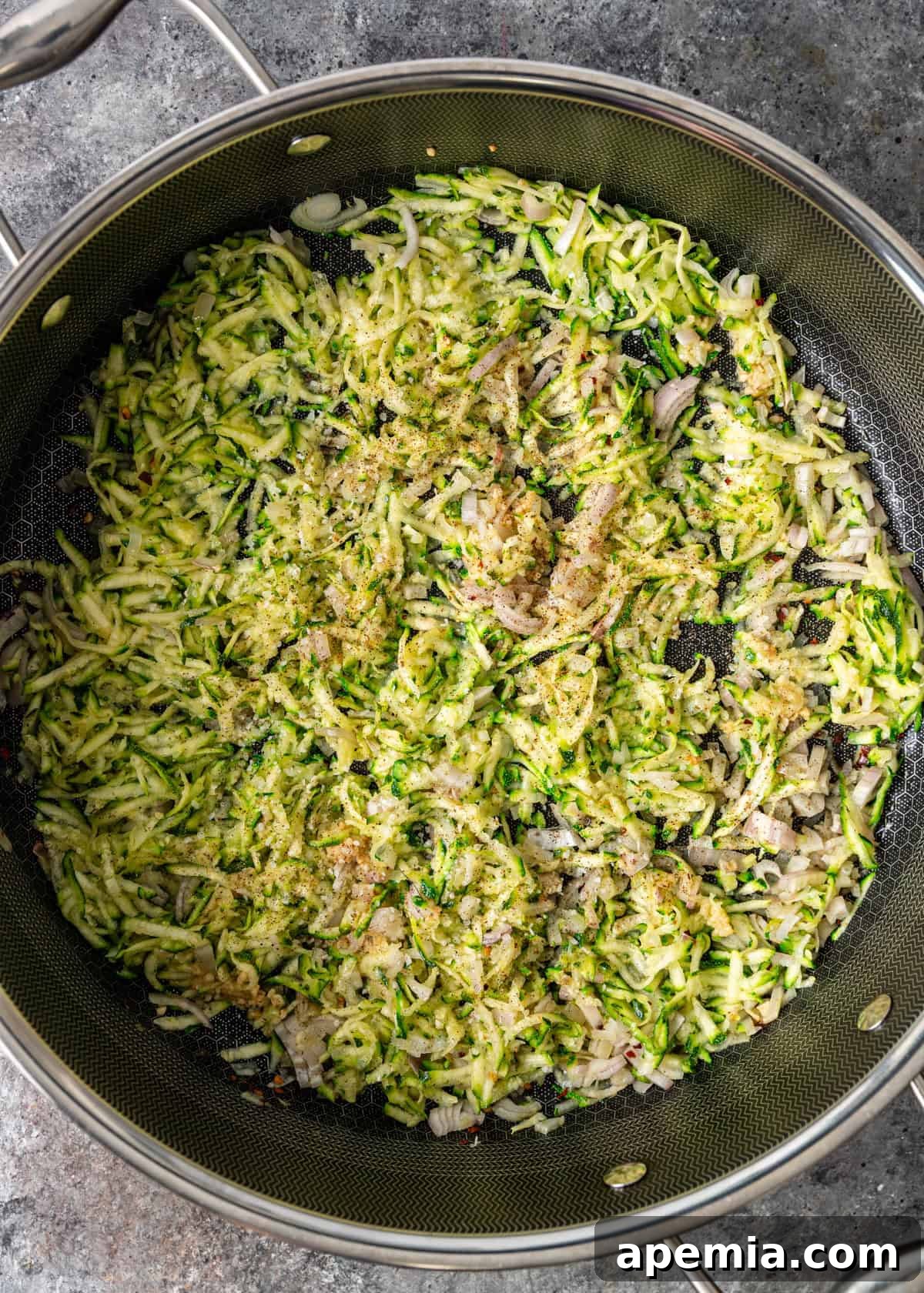 Overhead process shot: finely grated zucchini, thinly sliced shallots, and minced garlic beginning to cook in a skillet with olive oil