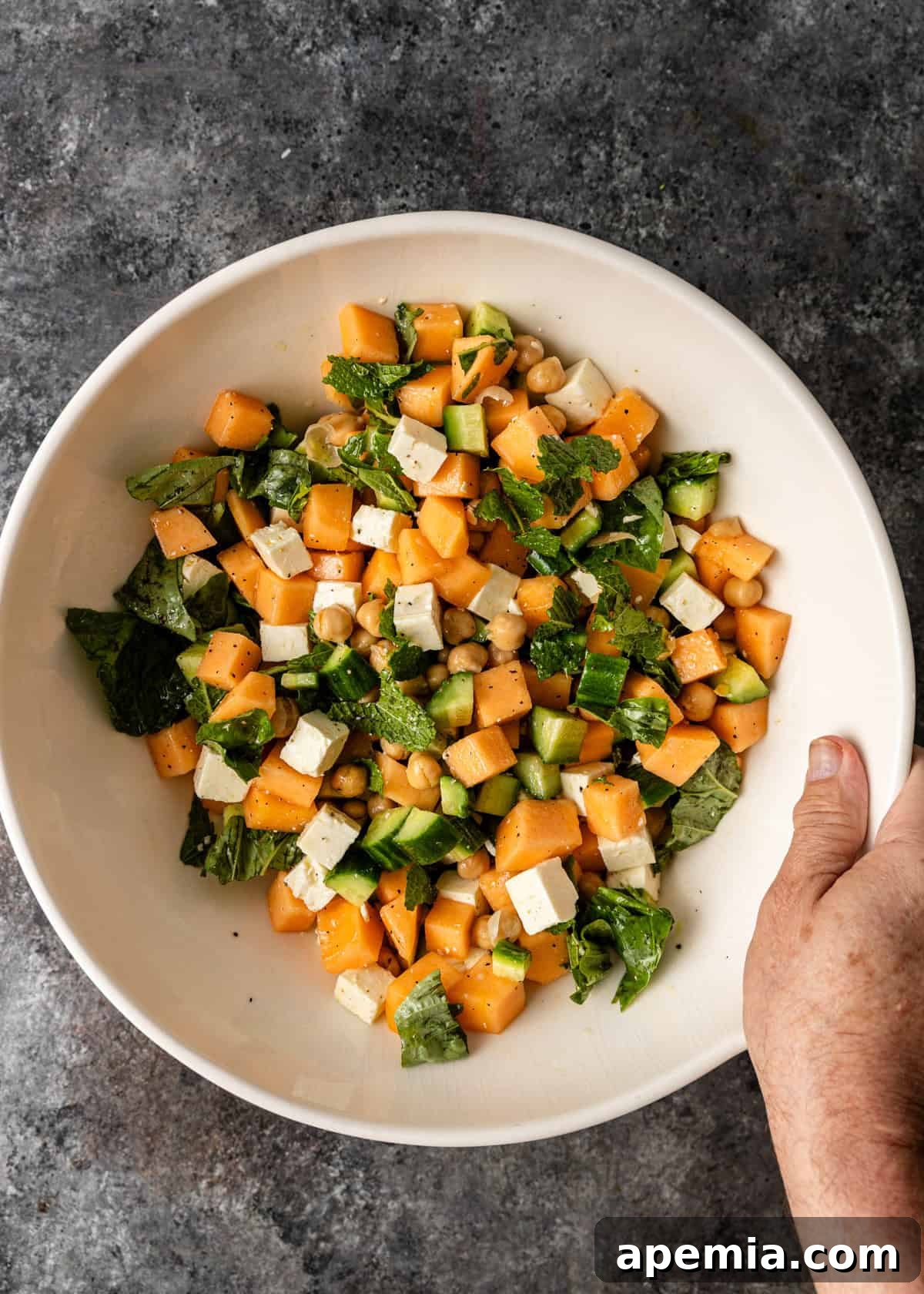 overhead: a hand holding a large white bowl of cantaloupe salad with feta and chickpeas