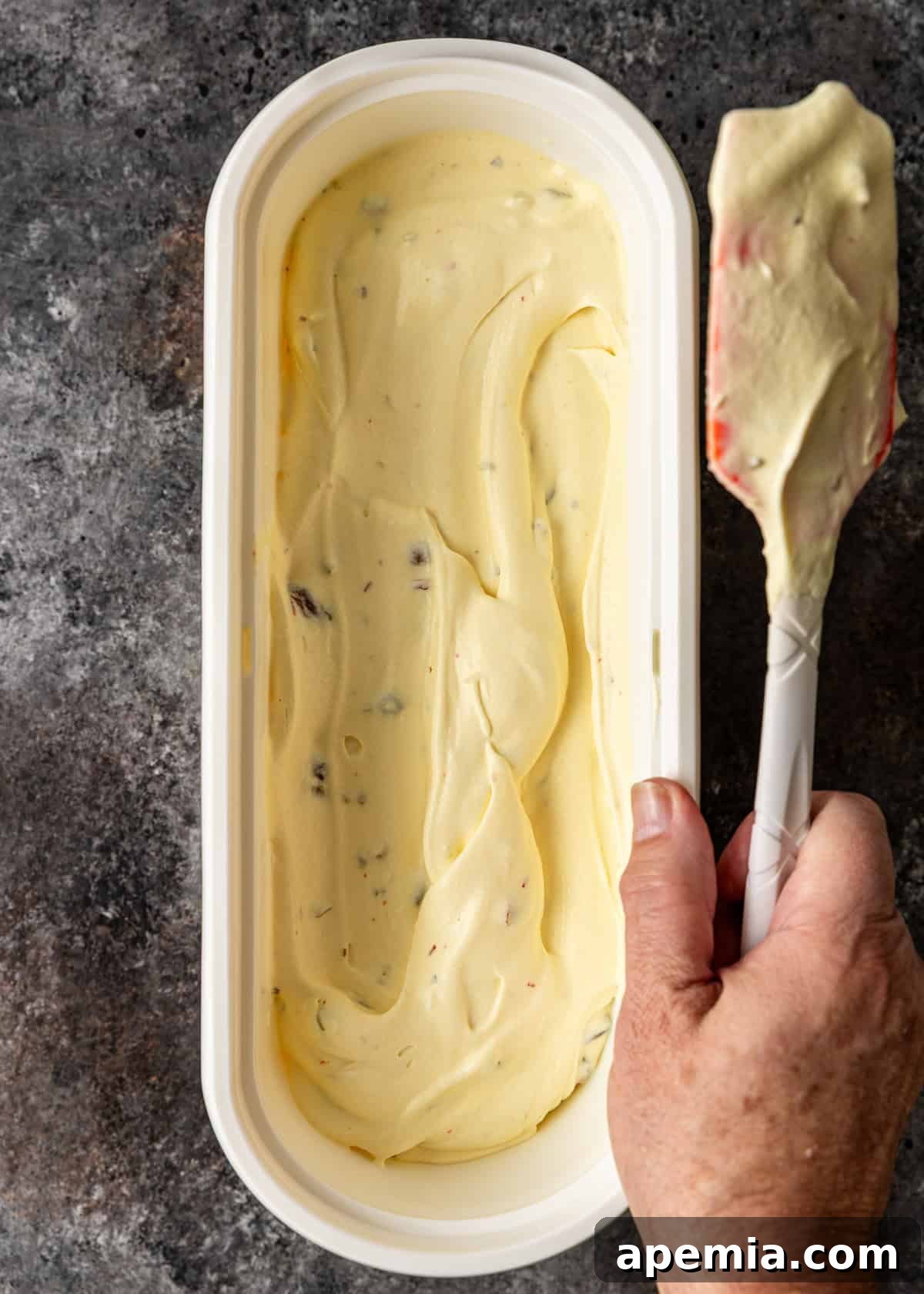 Overhead view: A hand using a spatula to smooth out the Bastani Sonnati ice cream within an insulated ice cream tub before freezing.