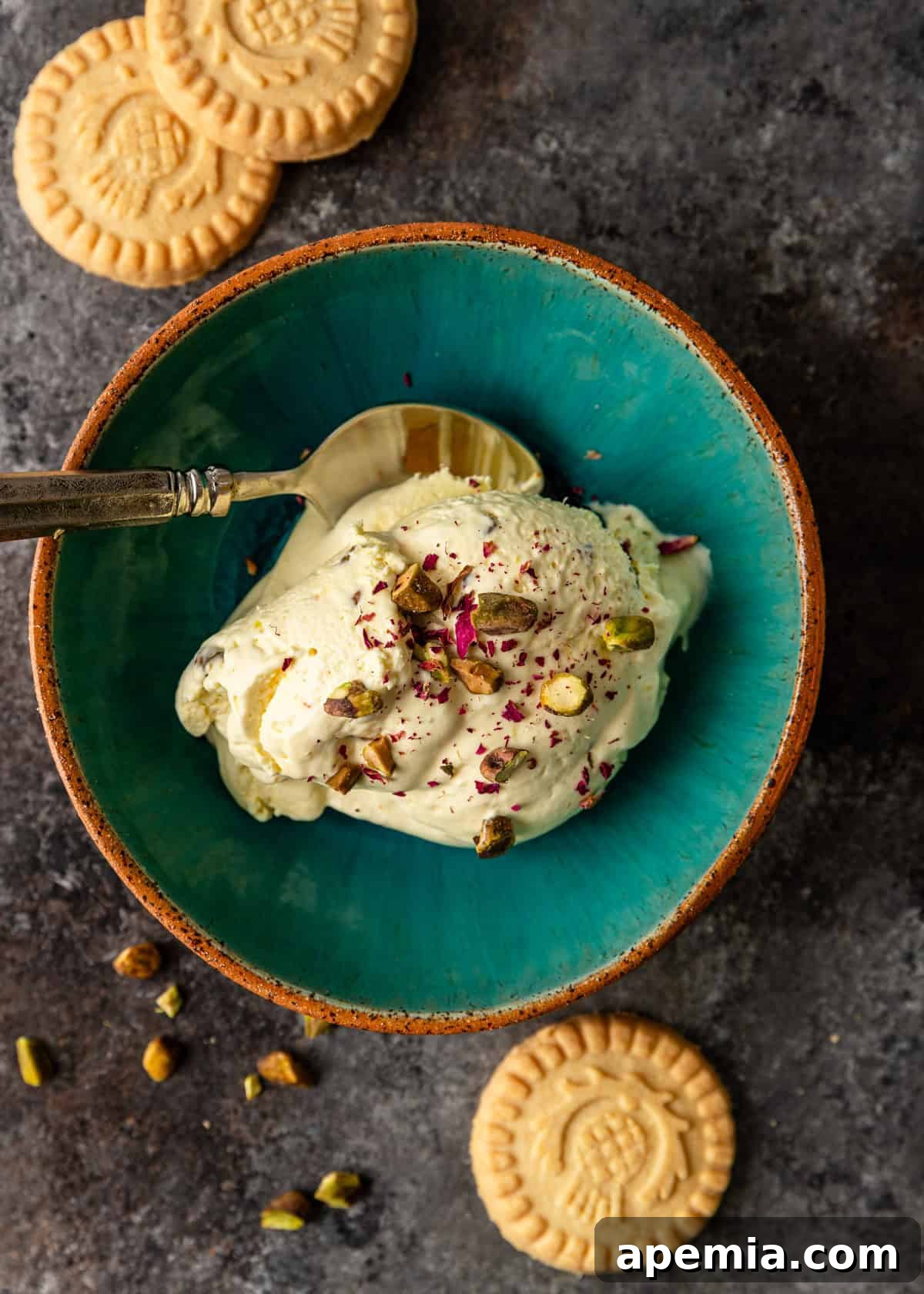 Overhead view: A few scoops of saffron and pistachio Persian ice cream, beautifully topped with crushed pistachios and fresh rose petals, served in an elegant blue bowl with a spoon.