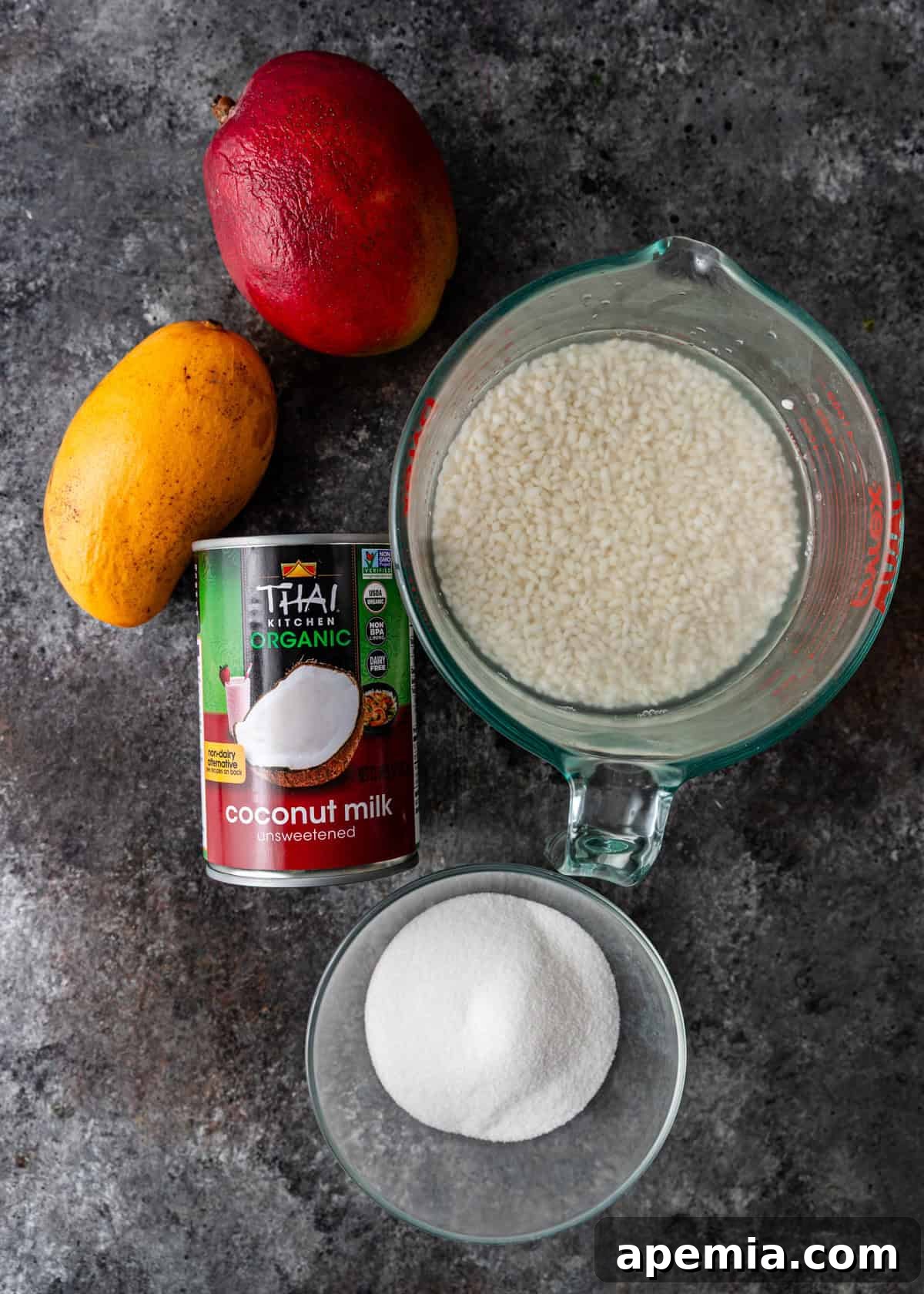 Overhead flat lay of key mango sticky rice ingredients: ripe mangoes, a can of coconut milk, uncooked sticky rice, and granulated sugar