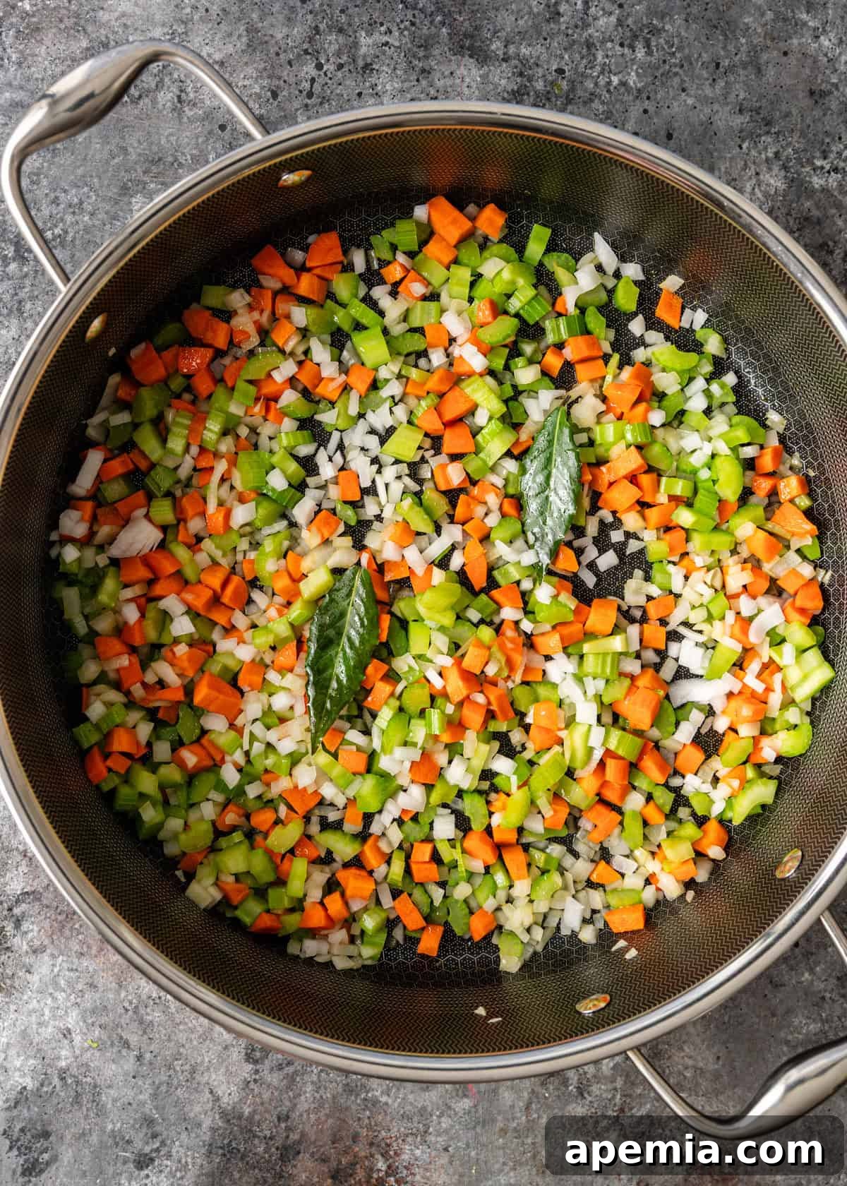 overhead process shot: large deep skillet on countertop with cut up celery, onion, carrot and bay leaves sautéing