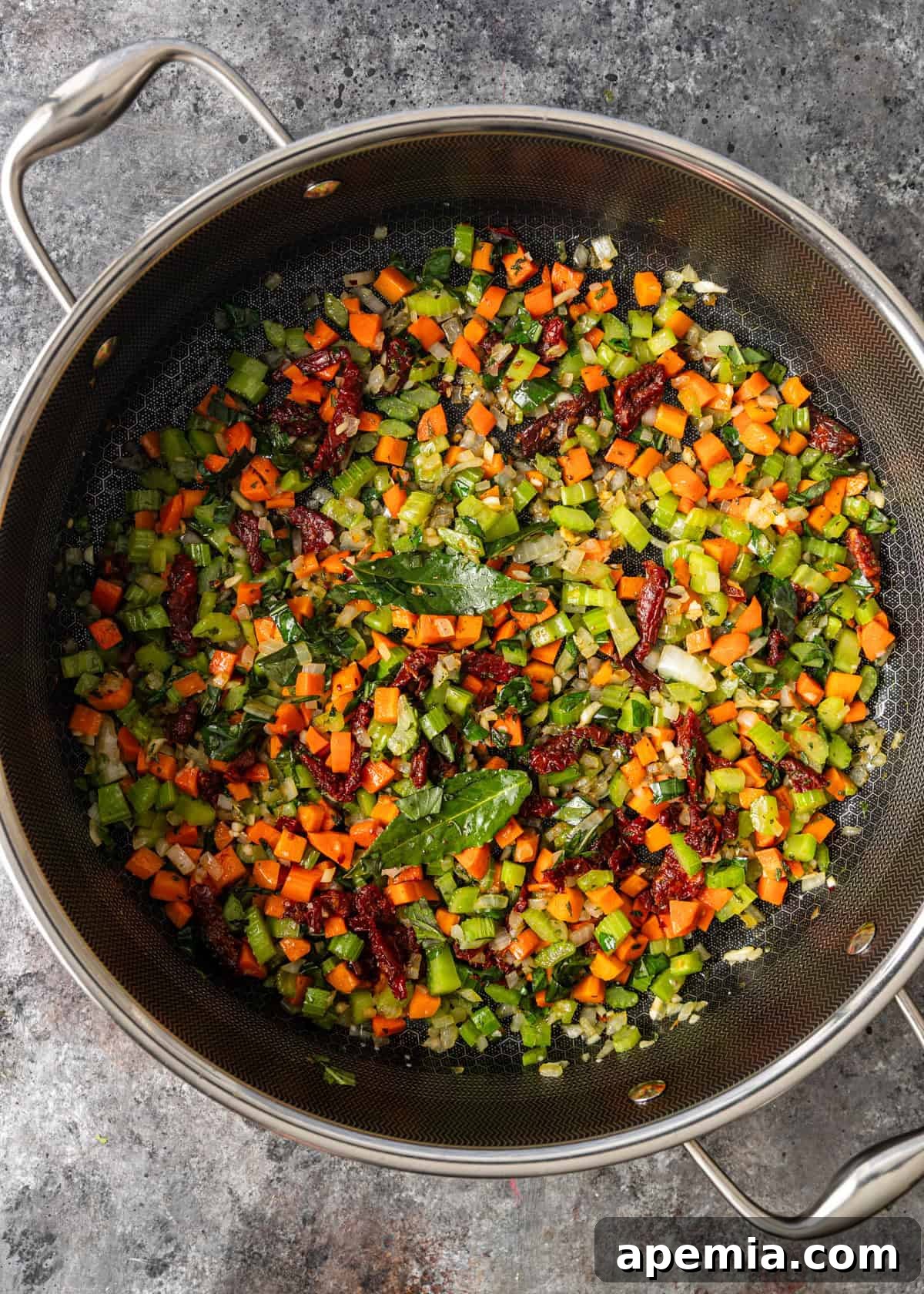overhead process shot: large deep skillet on countertop with cut up celery, onion, carrot, bay leaves, garlic, parsley, basil and sun-dried tomatoes added