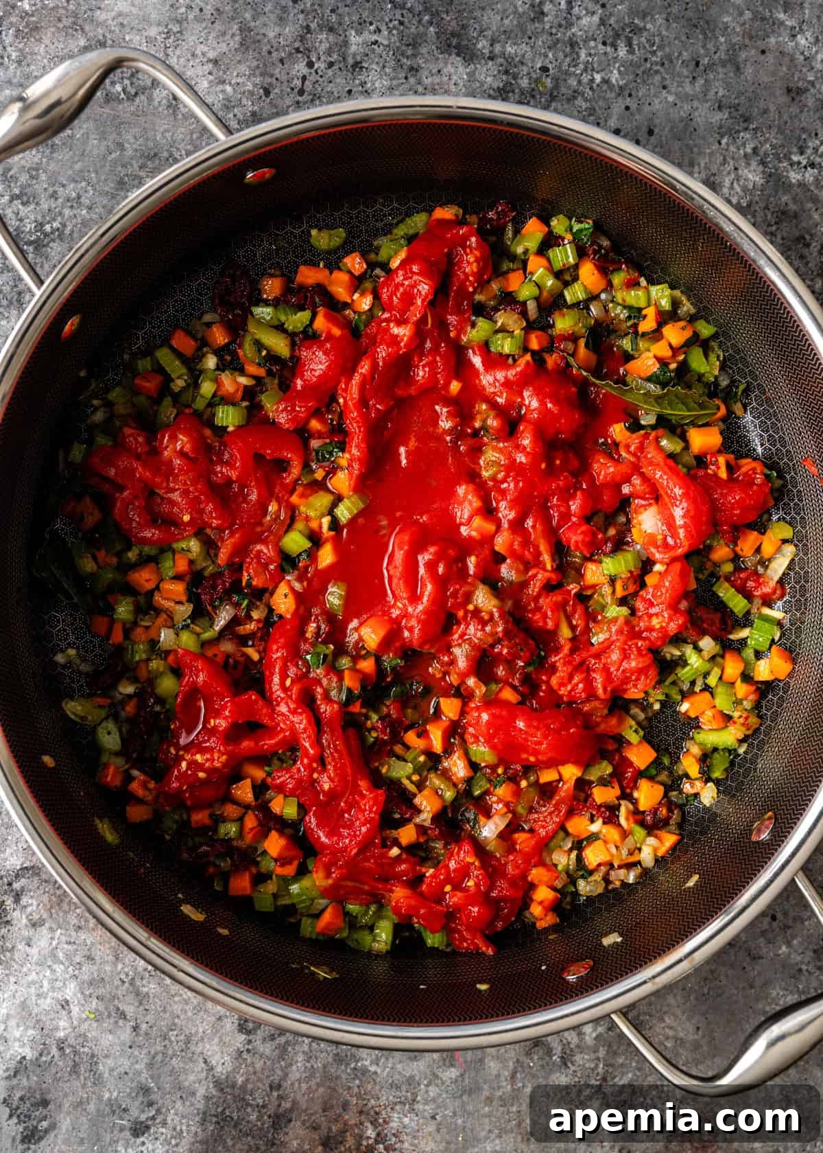 overhead process shot: large deep skillet on countertop with celery, onion, carrot, bay leaves, garlic, parsley, basil, sun-dried tomatoes, tomato paste, salt and red pepper flakes cooking down