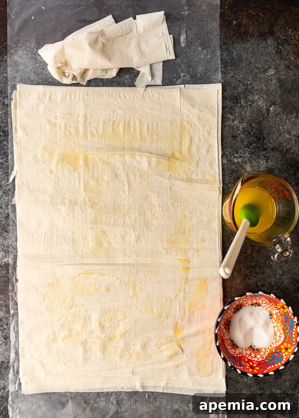 overhead process shot: phyllo dough on wax paper with measuring cup of butter off to side and bowl of sugar