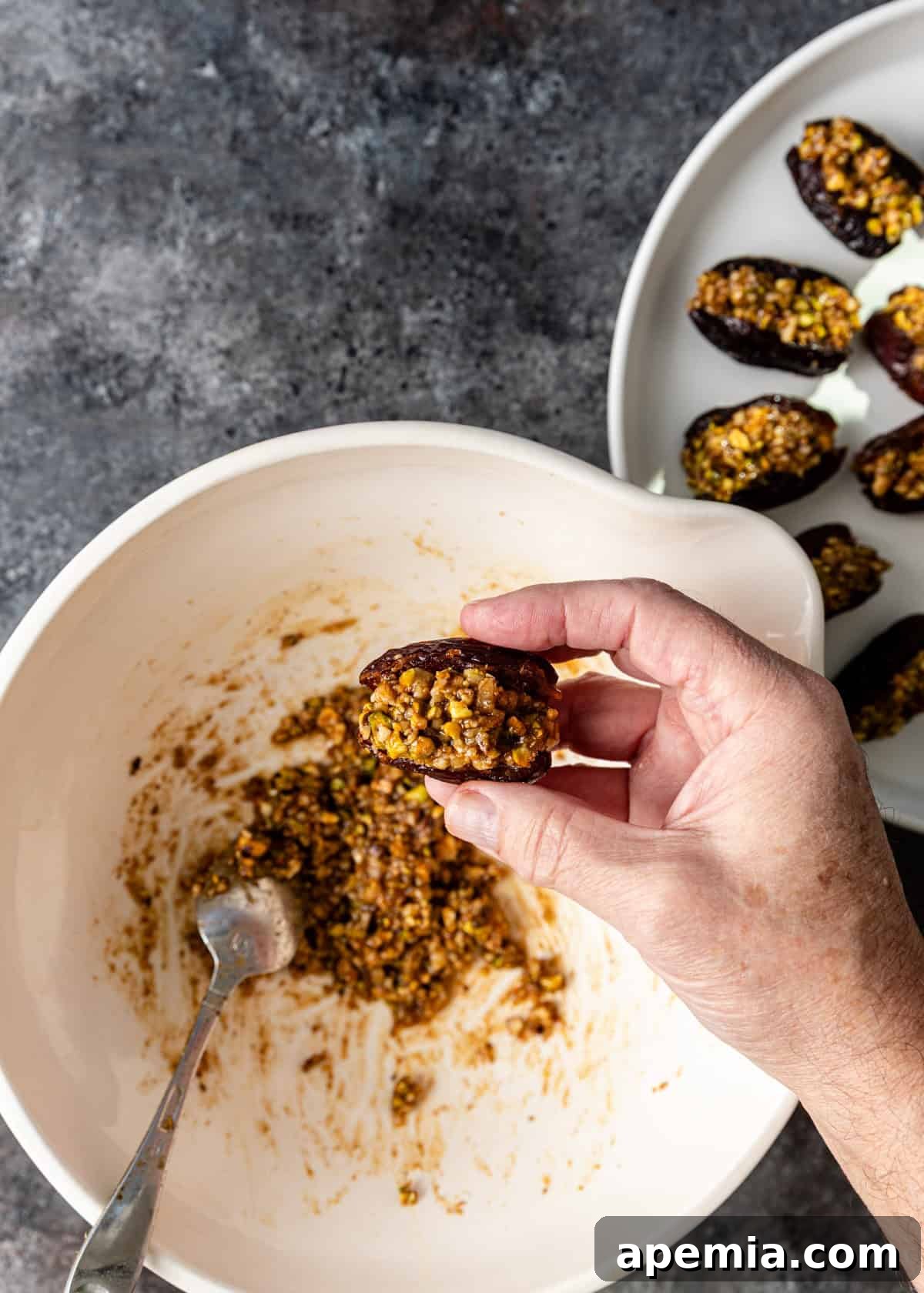 Overhead process shot: a collection of Baklava Stuffed Dates arranged on a plate, with one date being held above the bowl of remaining filling, showcasing the final product.