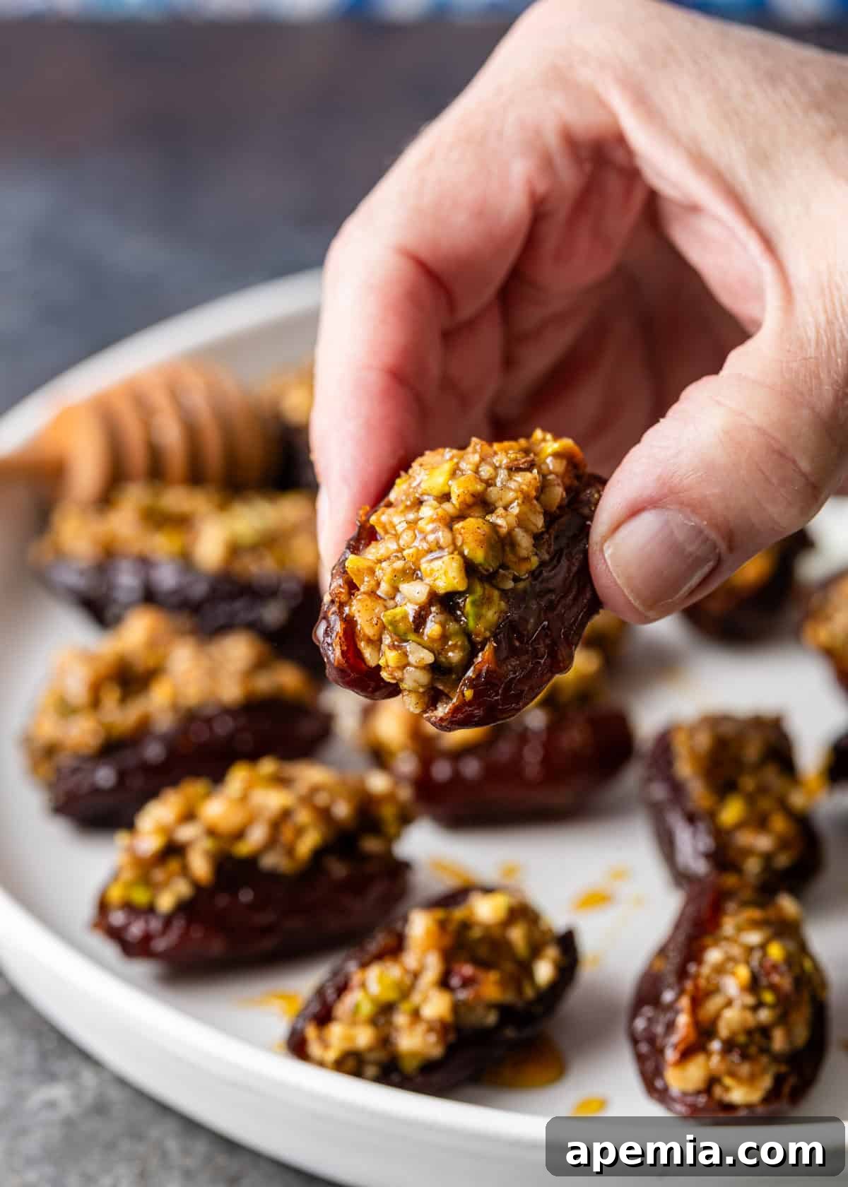 Closeup: a hand gently holding a single Baklava Stuffed Date over a white plate filled with more, showcasing the deliciousness ready to be enjoyed.