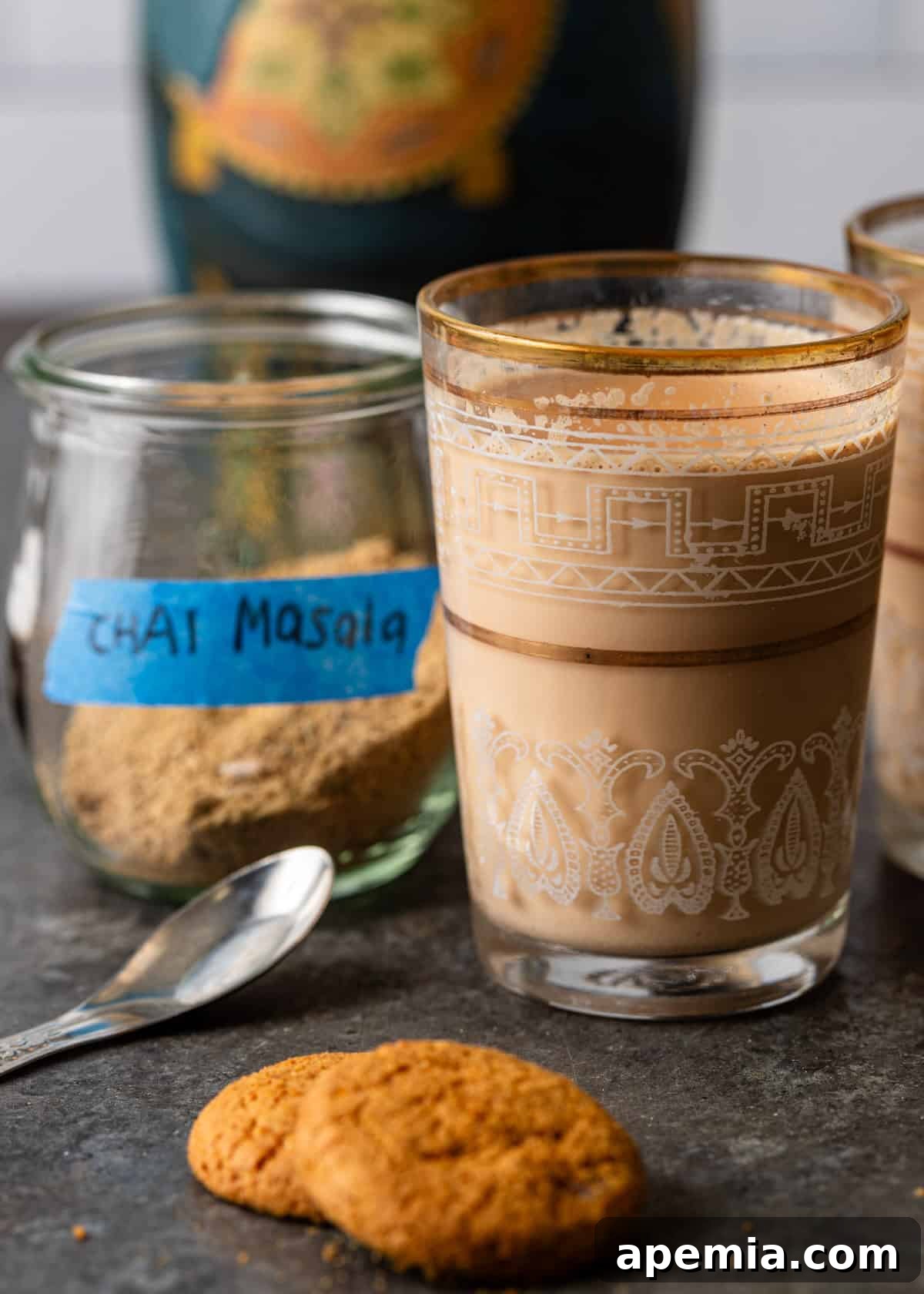 closeup side view: glass of chai in front of jar holding chai masala spices with spoon and two cookies on counter