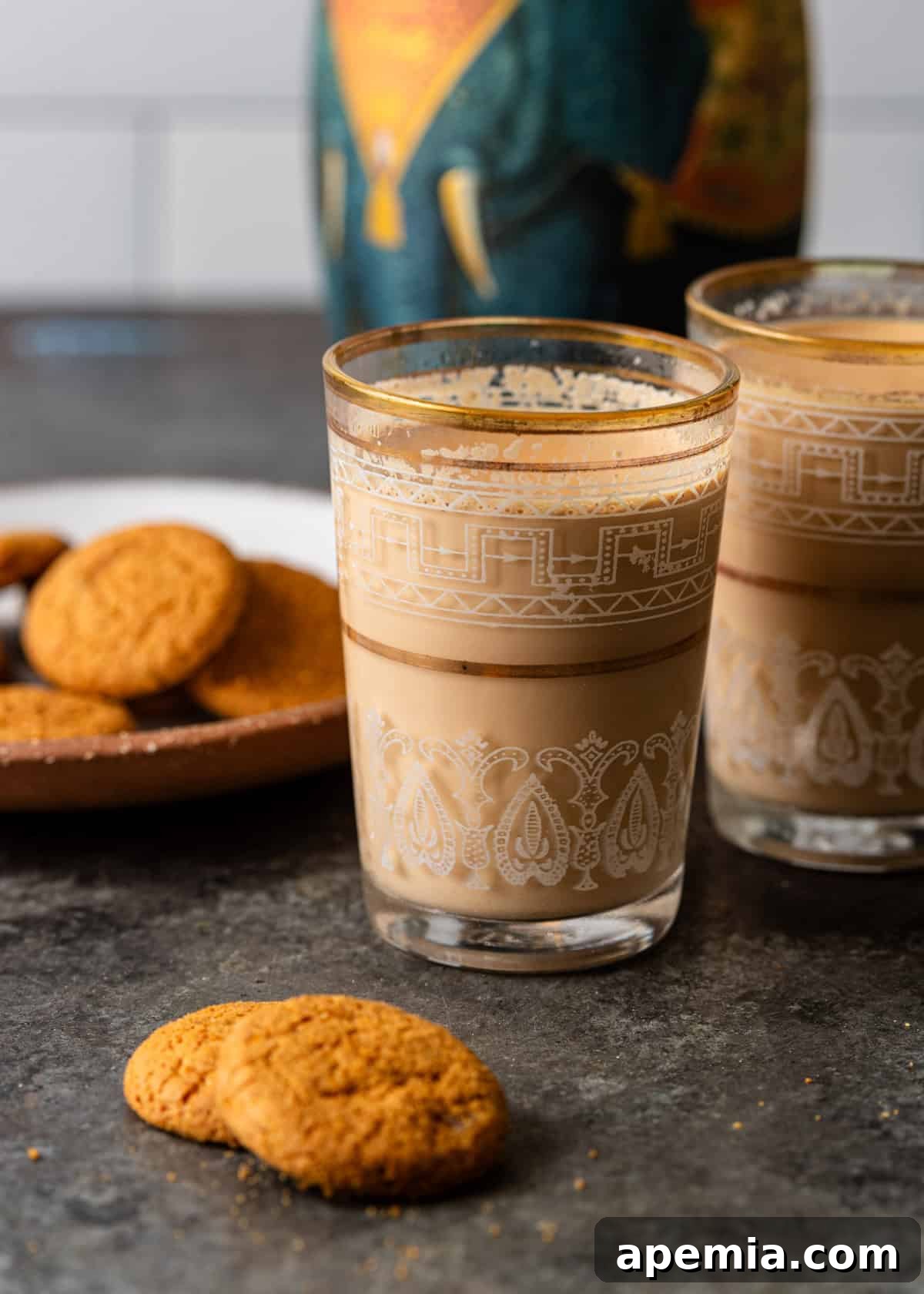 closeup side view: two glasses of masala chai with plate of cookies on plate and two cookies on counter in front of glasses