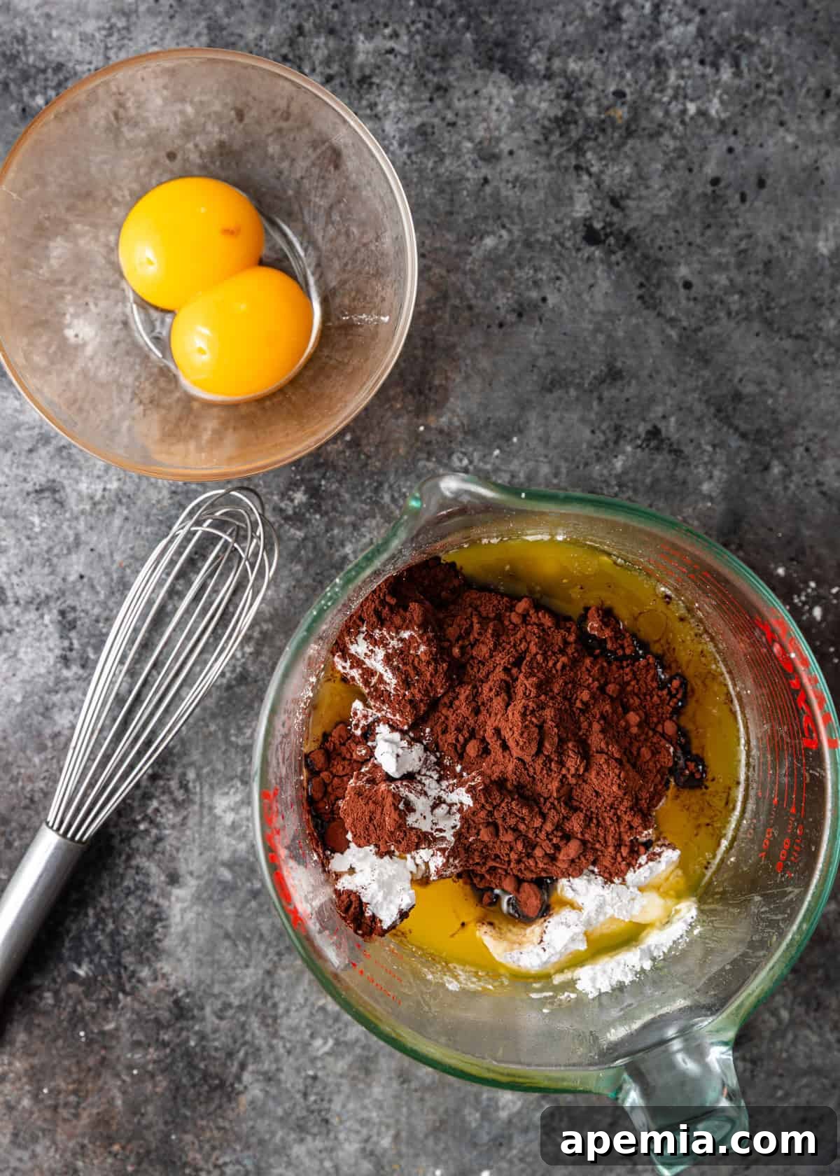 overhead process shot: whisk on counter with bowl of two egg yolks and measuring cup with chocolate mixture ingredients