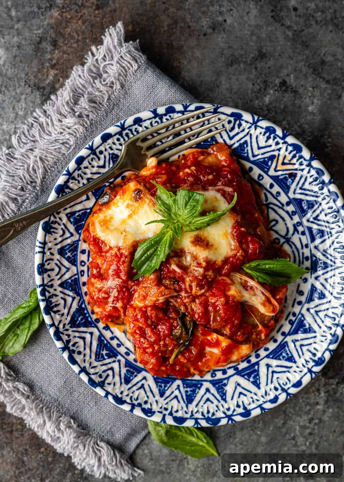 overhead: a big slice of parmigiana on white and blue plate topped with basil and fork on side