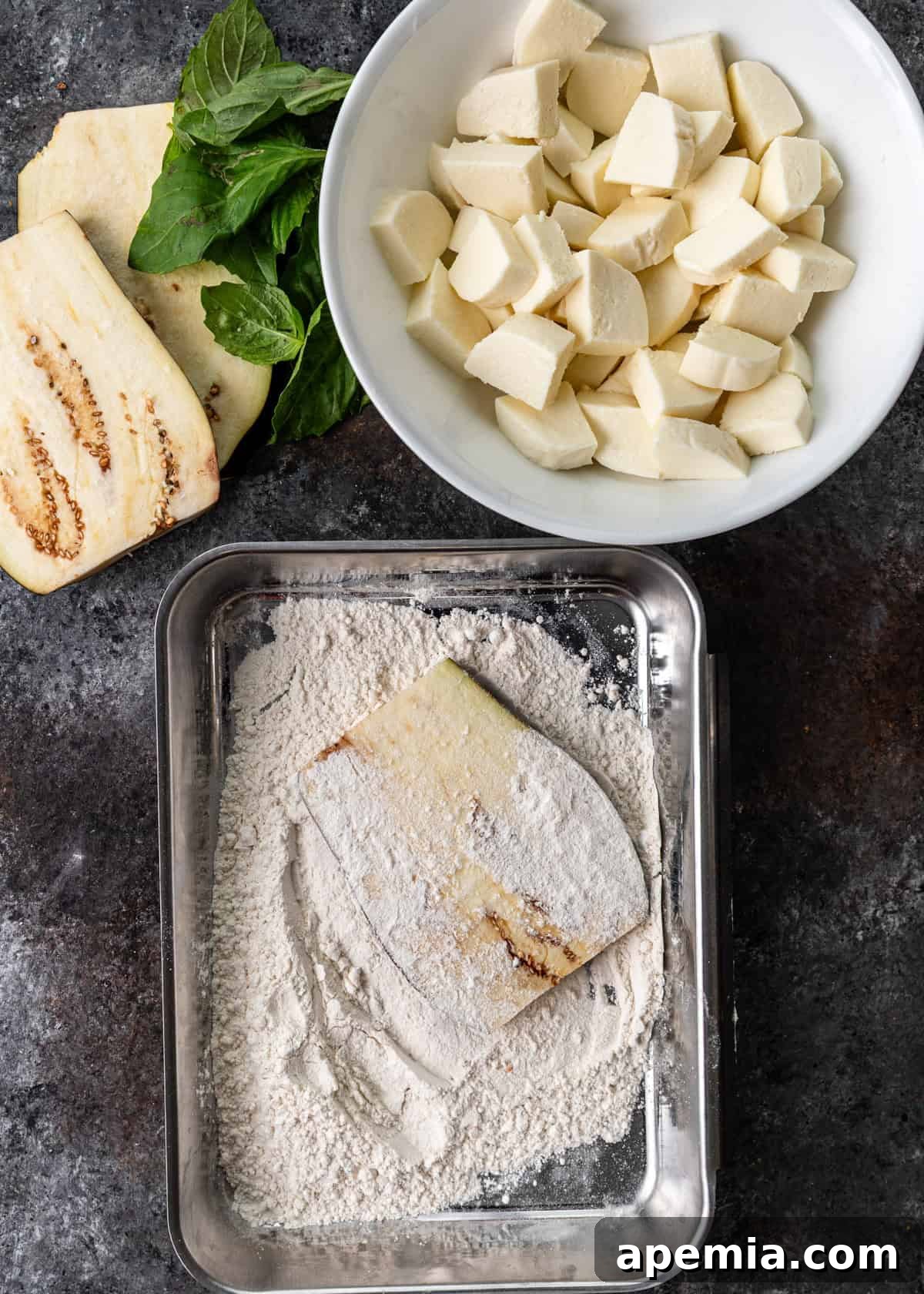overhead process shot: two slices of eggplant on counter with basil besides bowl of cut mozzarella and pan with flour covering a slice of eggplant