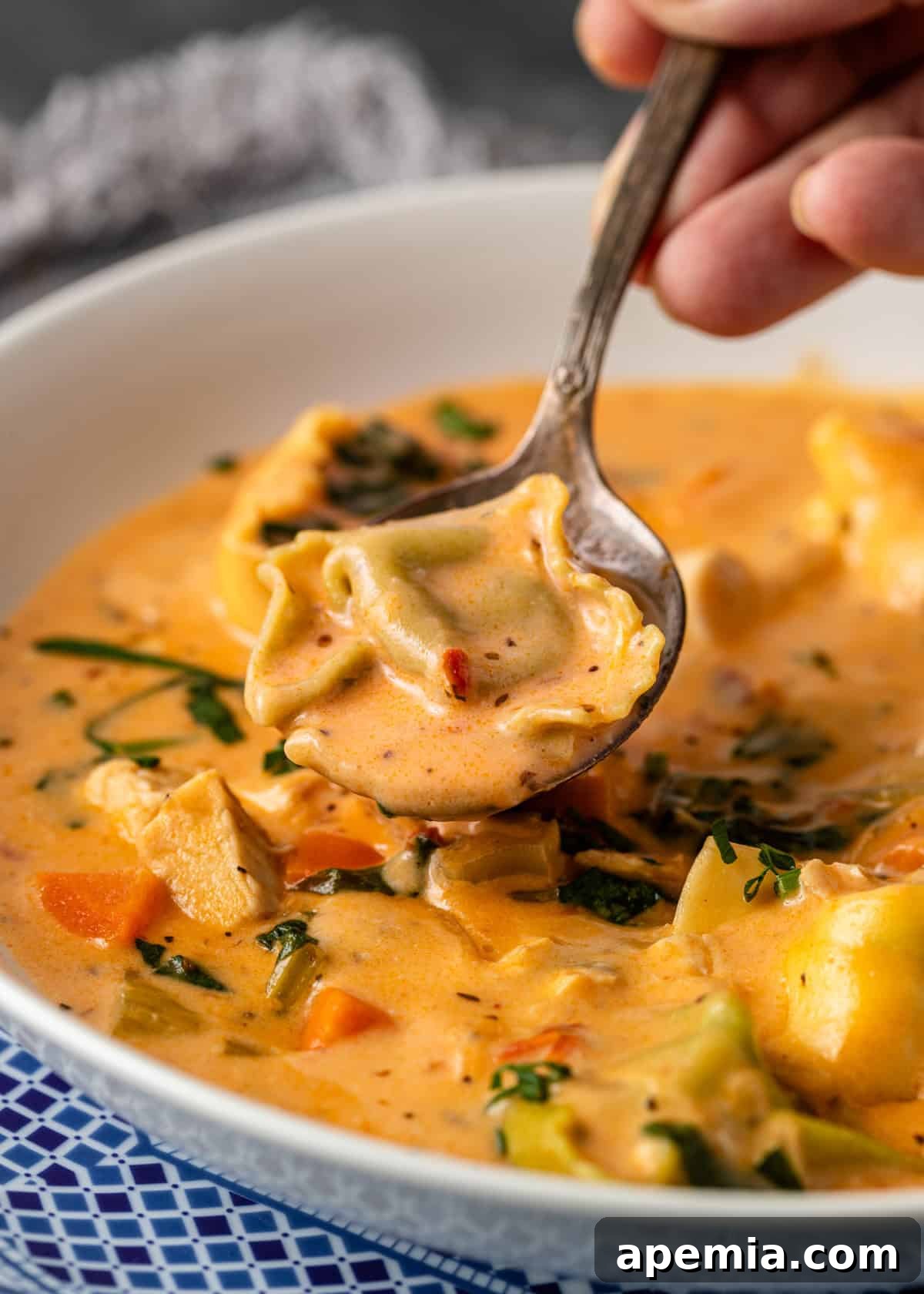 Close-up view of a hand holding a spoonful of creamy chicken tortellini soup above a white bowl, showcasing the rich texture and ingredients.