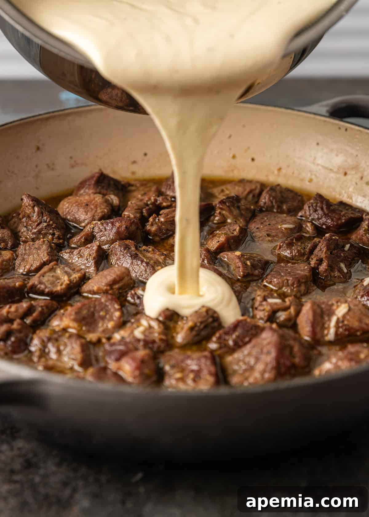 closeup side view: sauce being poured on top of cooked lamb cubes in skillet