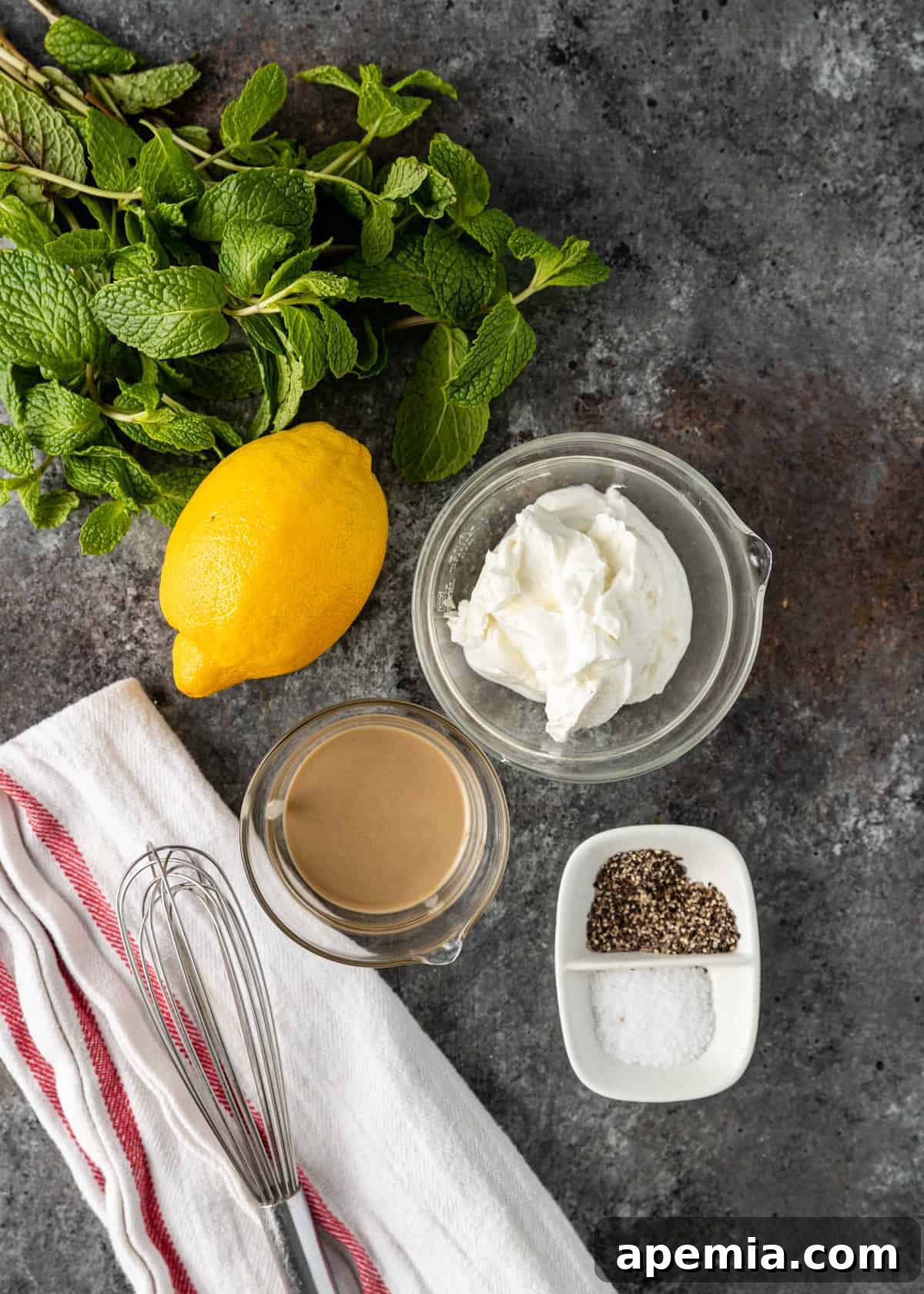 overhead image: bowl of Greek yogurt, whole fresh lemon, fresh mint, tahini paste, and salt and pepper, all on a gray backdrop with a whisk and kitchen towel