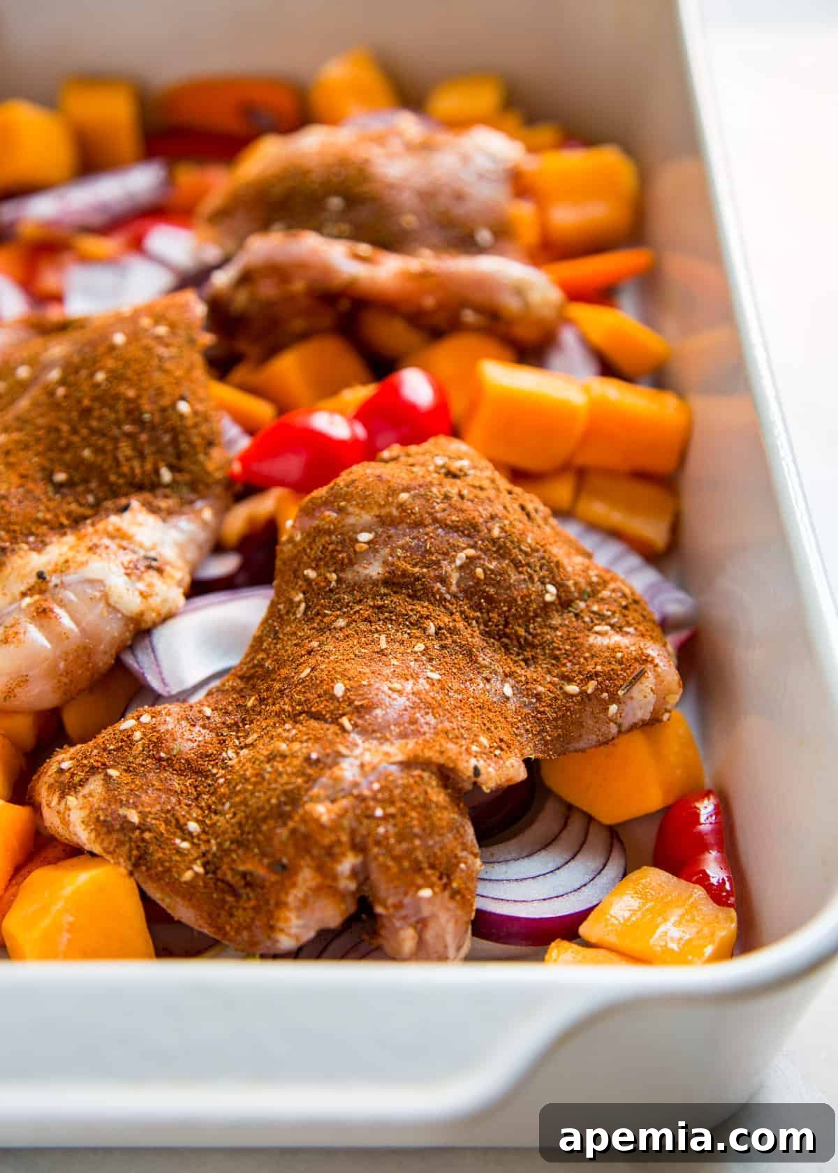 Raw chicken pieces arranged in a white baking dish with carrots, red onion, and bell peppers, all seasoned with African spices