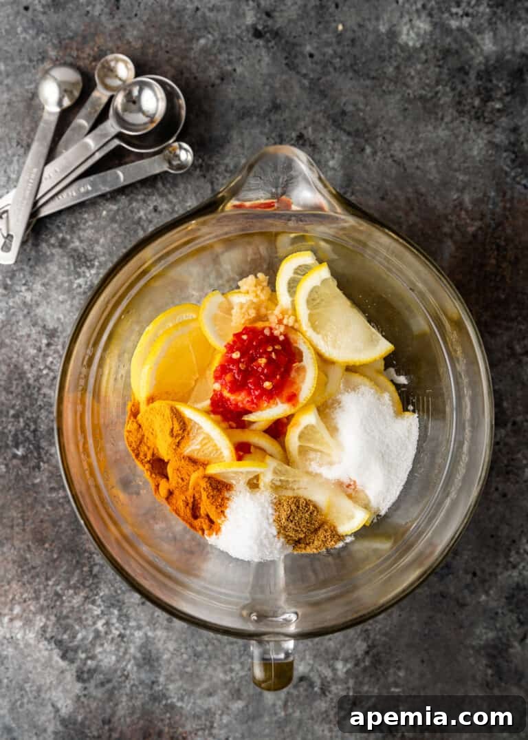 A glass mixing bowl filled with fresh lemon slices, ready for the pickling process, alongside various spices and a prepared chili paste.