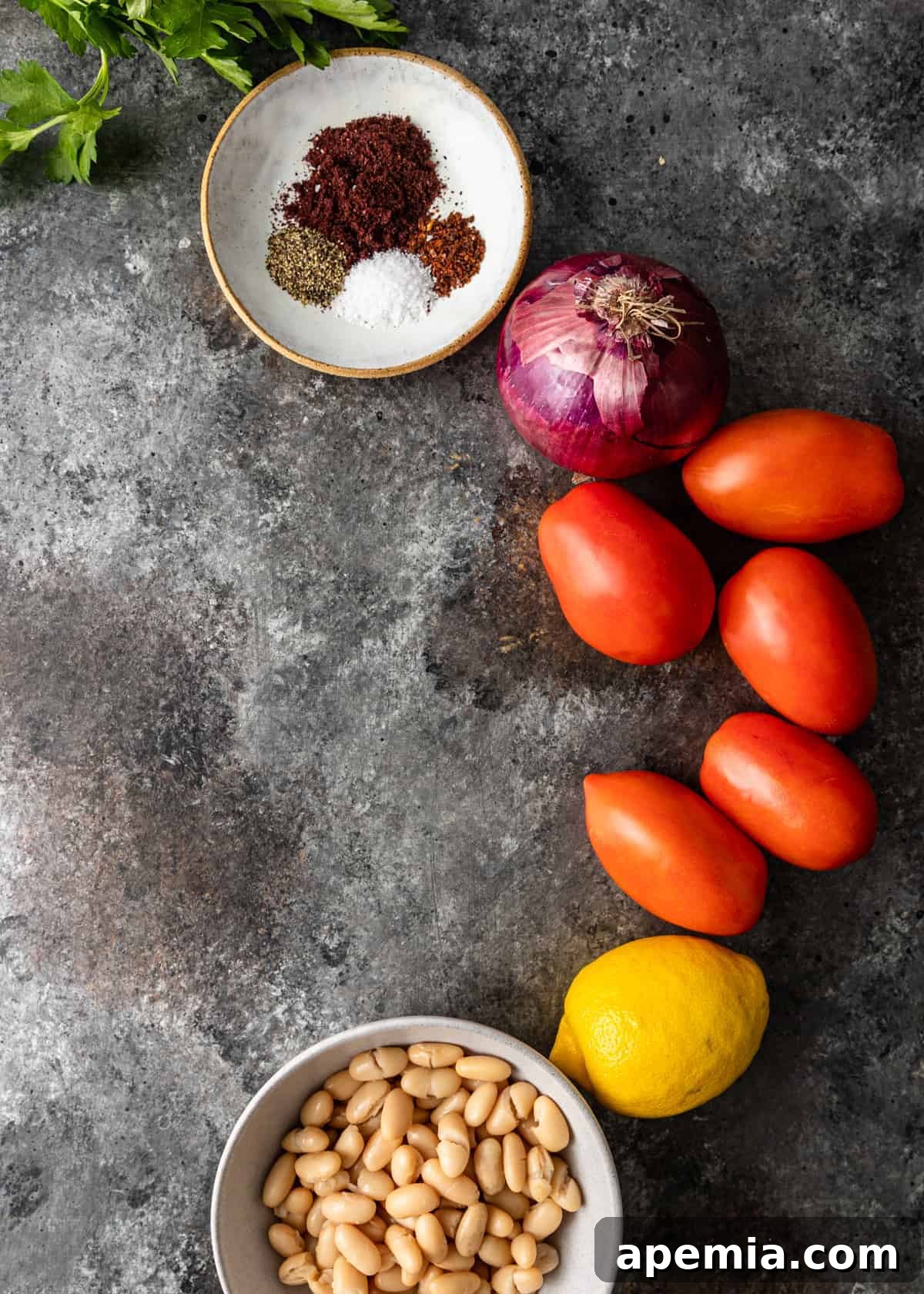 A top-down view displaying all the fresh, colorful ingredients required to make a delicious Turkish white bean salad, including white beans, tomatoes, red onion, parsley, lemon, olive oil, and spices.