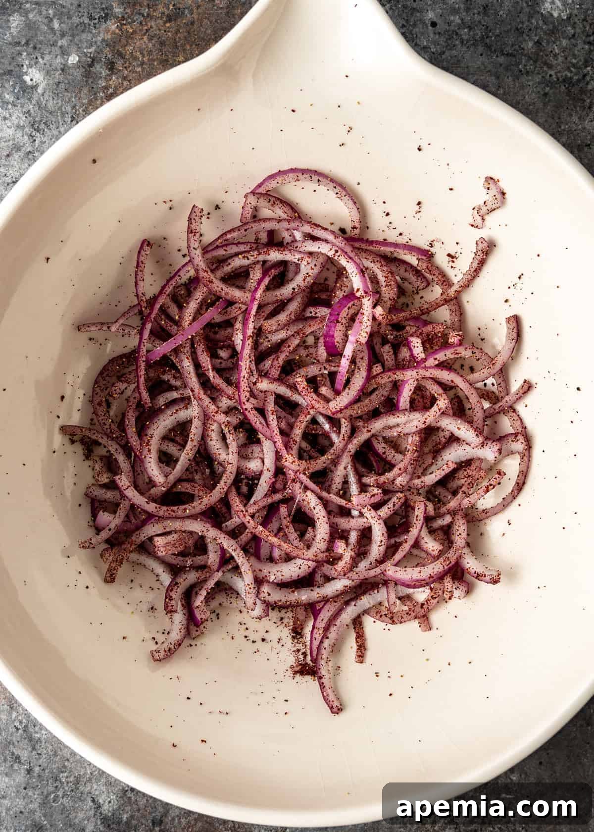 Close-up shot of thinly sliced red onions being massaged with sumac, Aleppo chili flakes, salt, and pepper in a white bowl, preparing them to soften for the Piyaz salad.