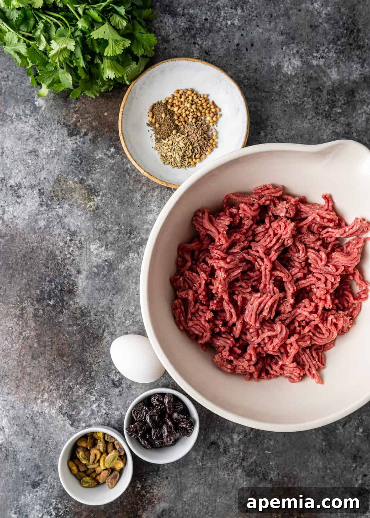 Overhead flat lay of individual ingredients including ground lamb, vibrant dried cherries, finely chopped pistachios, a fresh egg, and a selection of aromatic spices, all laid out for making Persian lamb meatballs.