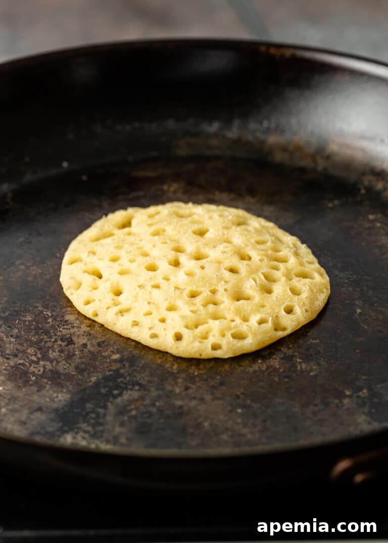 An overhead shot of a Baghrir pancake finishing its cook in a non-stick skillet, its surface completely transformed into a porous, honeycomb-like texture.