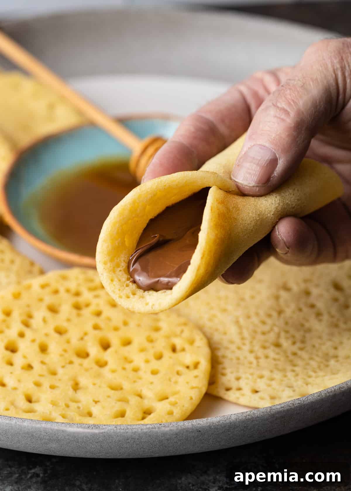 Close-up of a hand holding a golden-brown Baghrir pancake, folded and filled with a rich spread of Nutella chocolate hazelnut spread, ready to be eaten.