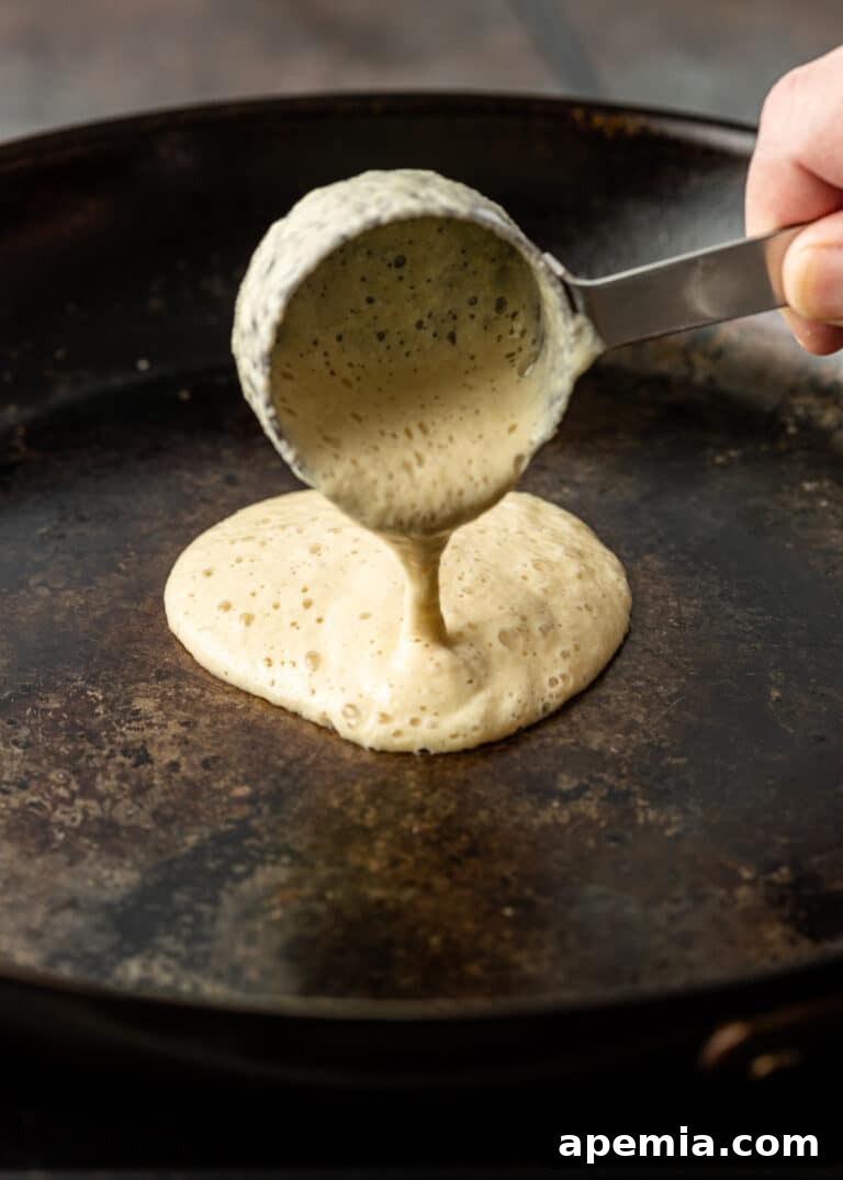 A stream of light yellow Baghrir batter being poured from a measuring cup into a hot non-stick skillet, indicating the start of the cooking process.