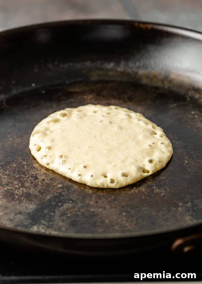 The cooking process of a Baghrir pancake in a skillet, with many tiny holes already formed on the surface, showcasing its distinctive honeycomb texture.