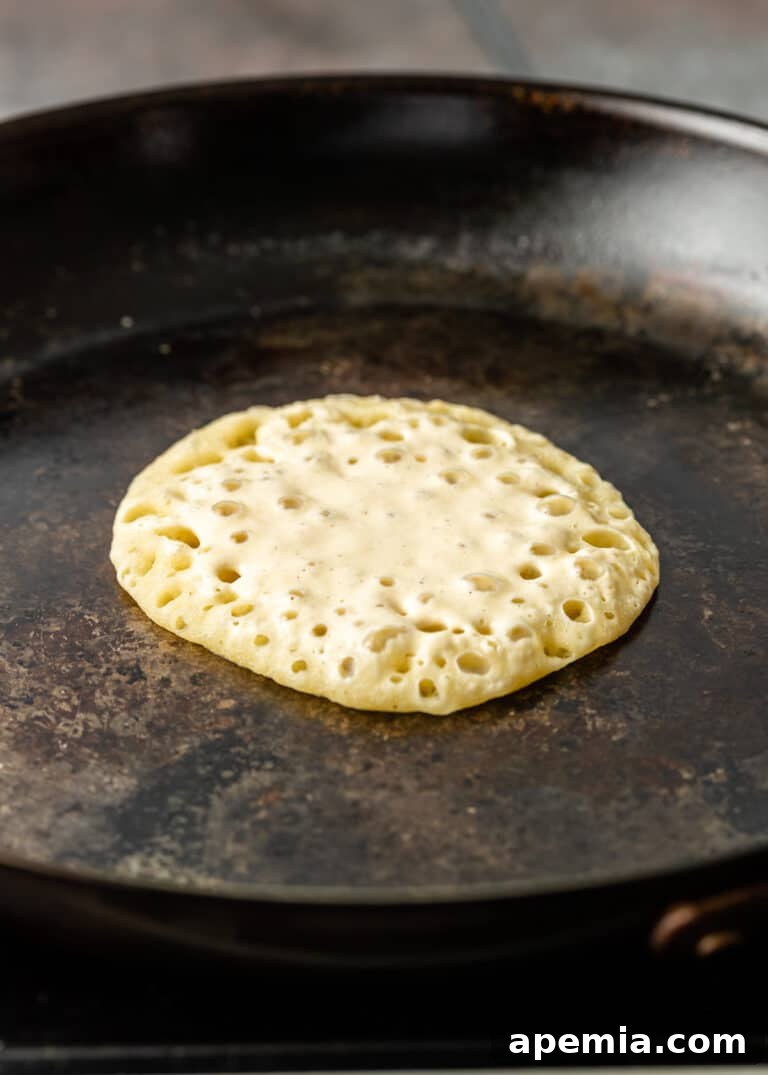 A detailed view of a Baghrir pancake in a skillet, with an intricate pattern of small, uniform holes spread across its entire surface, indicating it's nearly done.