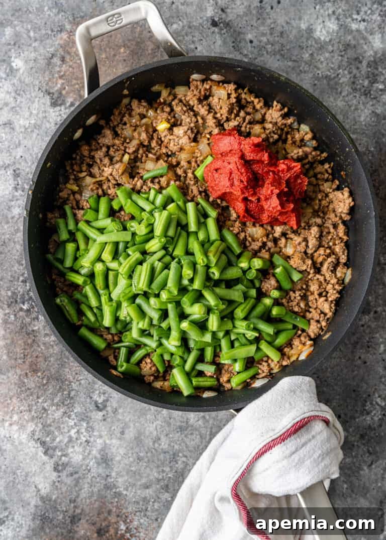 overhead process shot: ground lamb, green beans, and tomato paste in large skillet